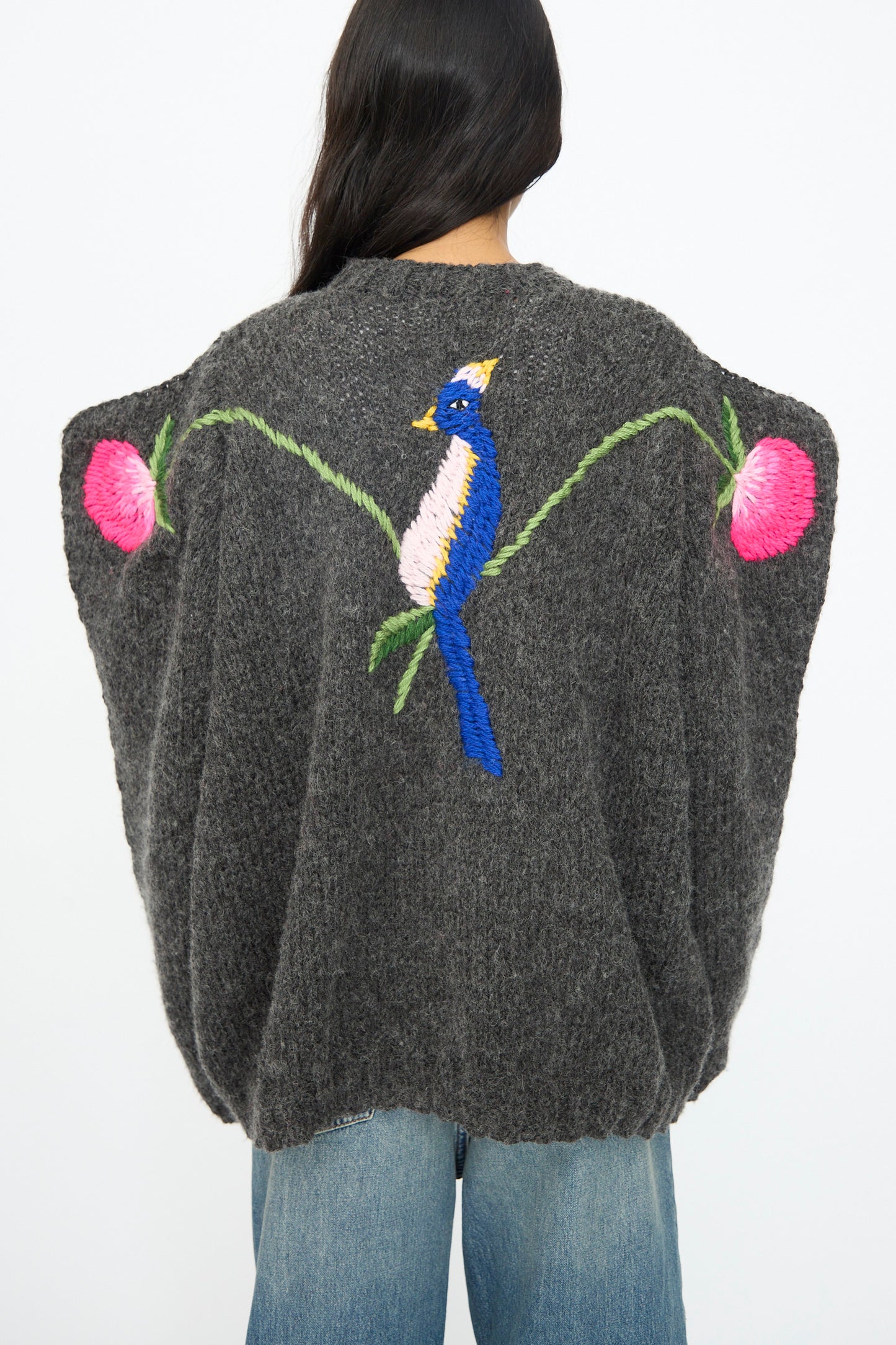 A person wears the Sofio Gongli Sweater in Charcoal with Birds and Flowers, featuring hand embroidery on the back, paired with blue jeans.