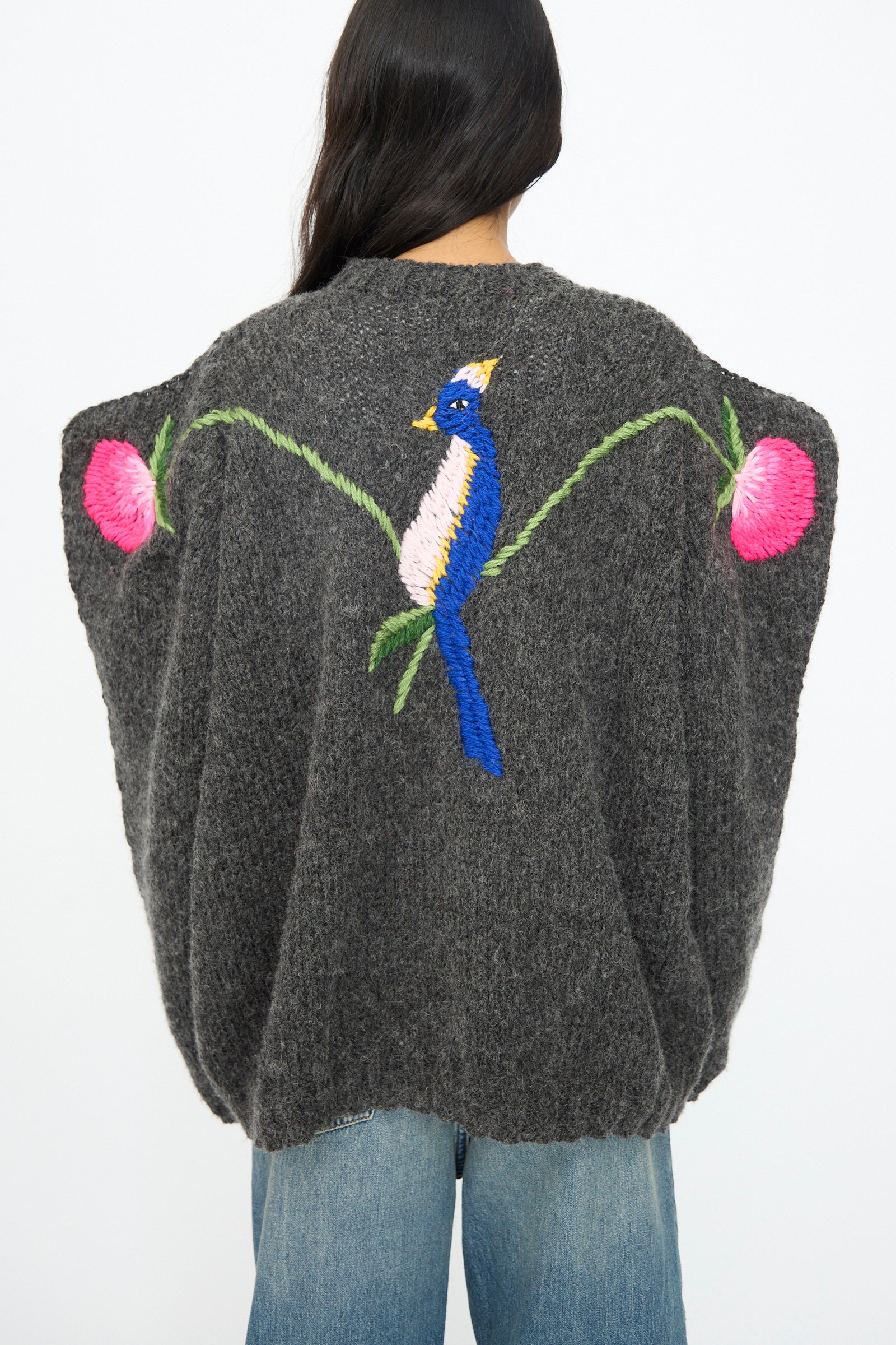 A person wears the Sofio Gongli Sweater in Charcoal with Birds and Flowers, featuring hand embroidery on the back, paired with blue jeans.