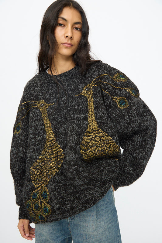 A woman wears the Sofio Gongli Sweater in Charcoal with Peacocks—a hand-knit mohair wool blend featuring two large gold peacock designs—paired with light blue jeans, against a plain background.