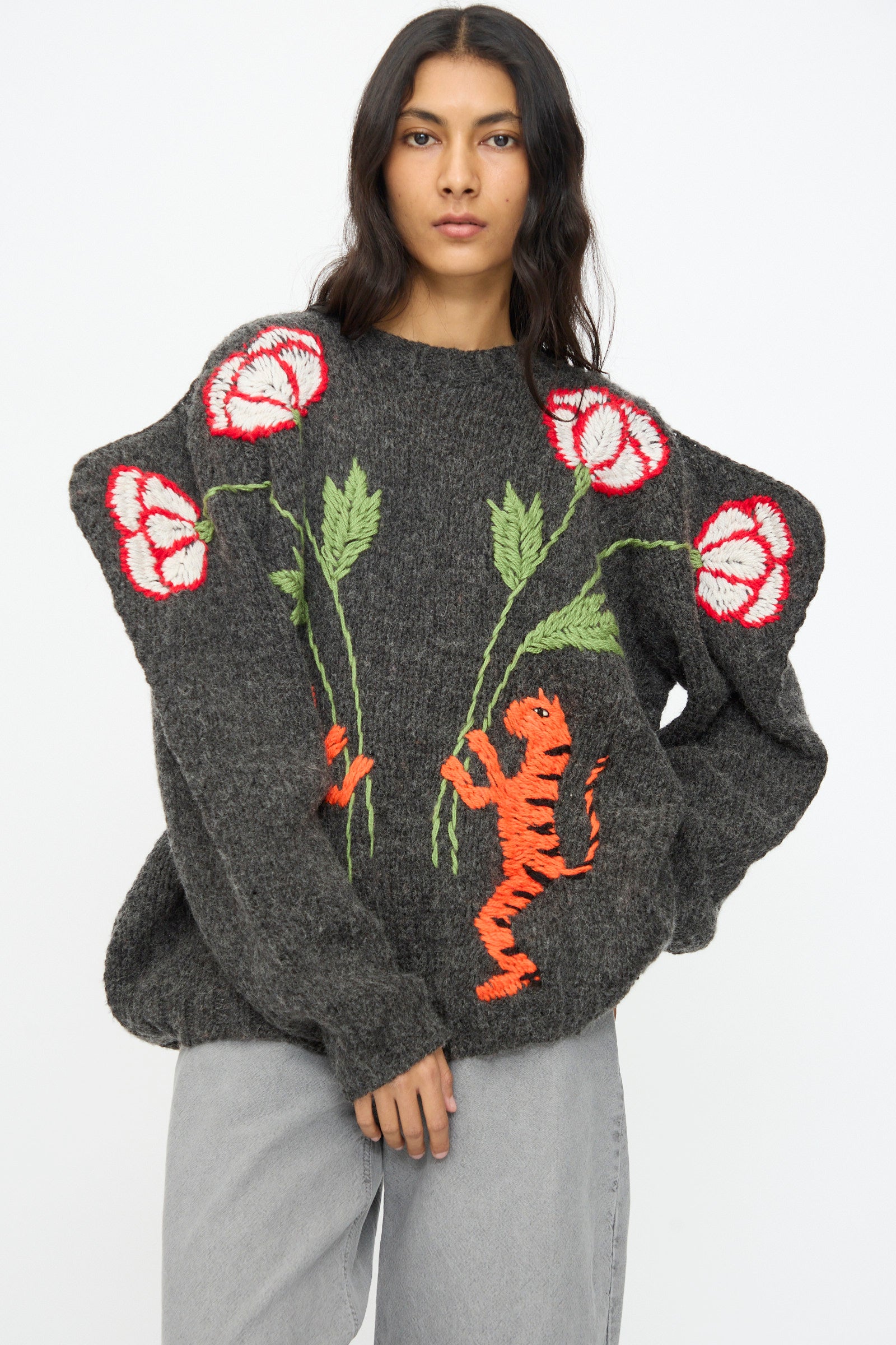 A woman wears the Sofio Gongli Sweater in Charcoal with Tigers and Flowers, an oversized mohair wool blend featuring red and white floral and orange tiger embroidery, paired with light gray pants against a plain background.