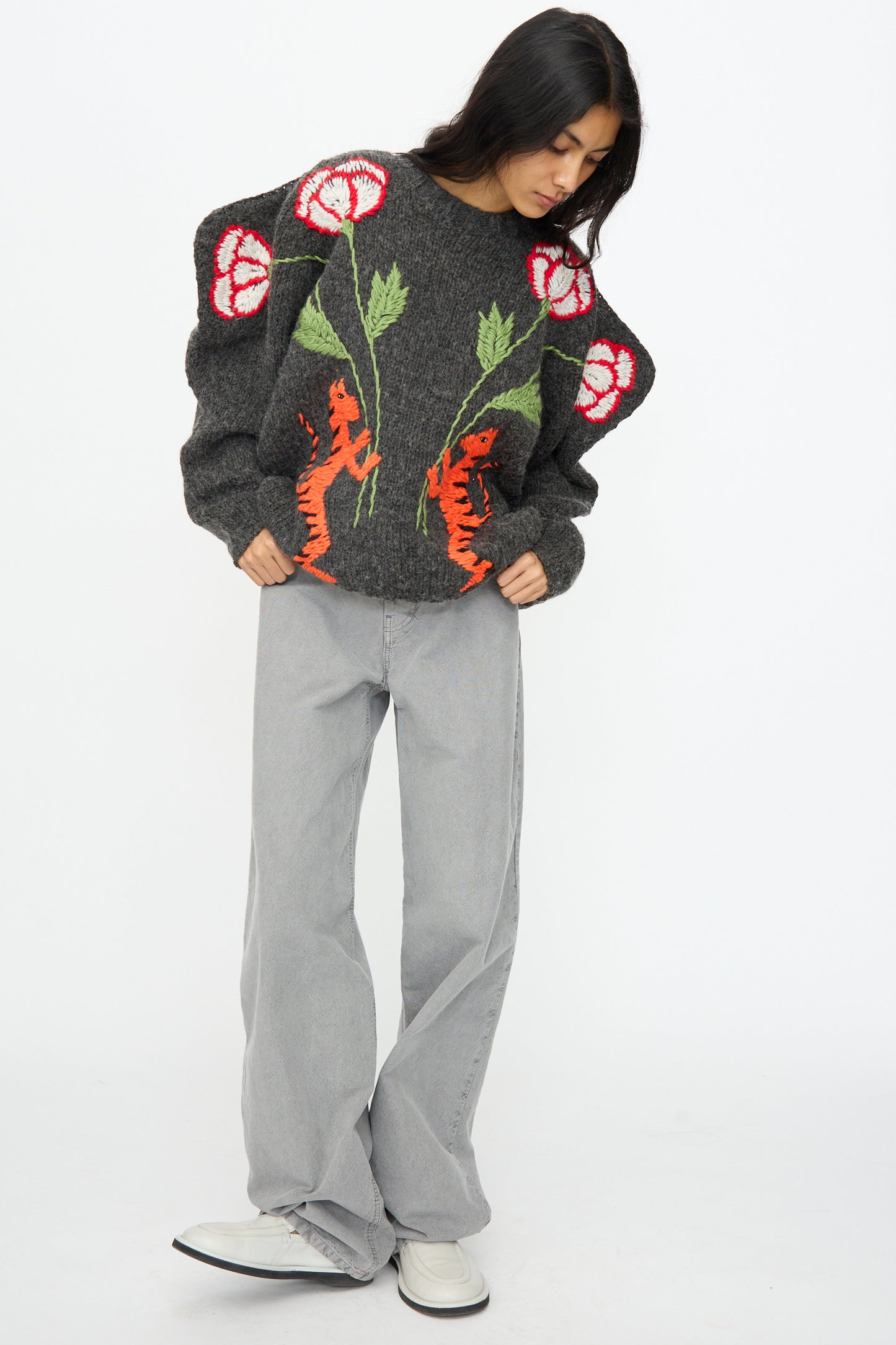 A woman wears the Sofio Gongli Sweater in Charcoal with Tigers and Flowers, paired with light gray wide-leg jeans and white shoes, standing against a plain white background.