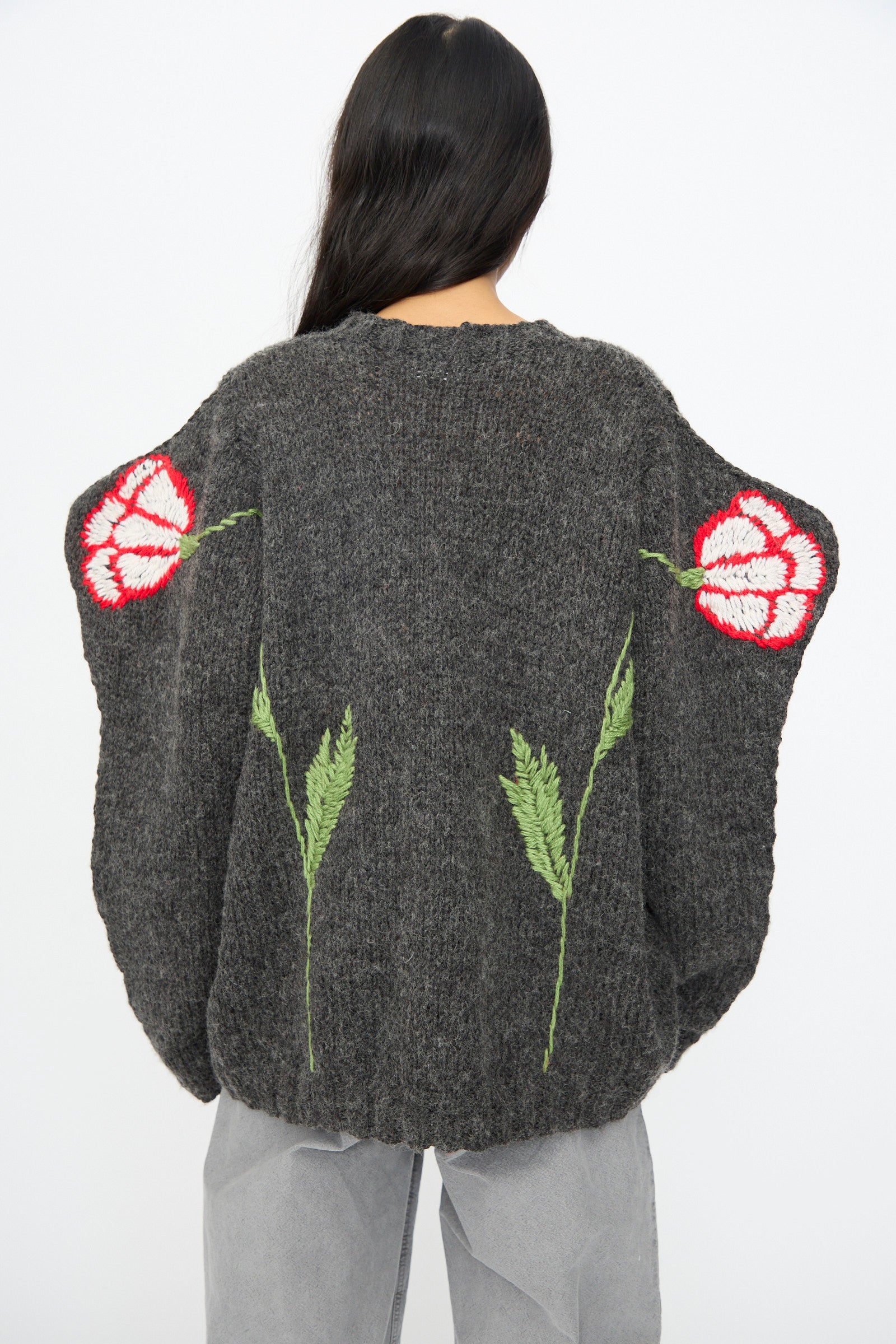 A person with long dark hair wears the Sofio Gongli Sweater in Charcoal with Tigers and Flowers, a loose-fitting hand-knit piece, styled with light gray pants.
