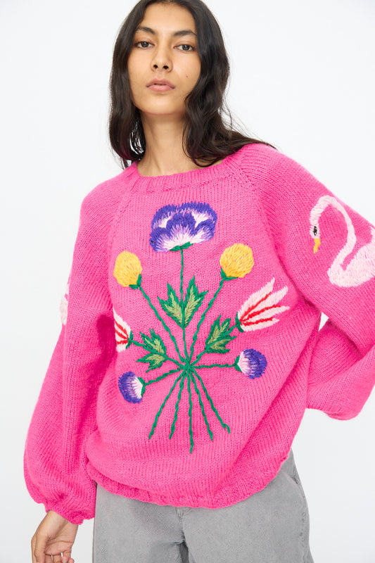 A person wears the Sofio Gongli Sweater in Pink with Flowers, featuring colorful embroidery and a flamingo sleeve design in soft mohair blend, paired with light gray pants.