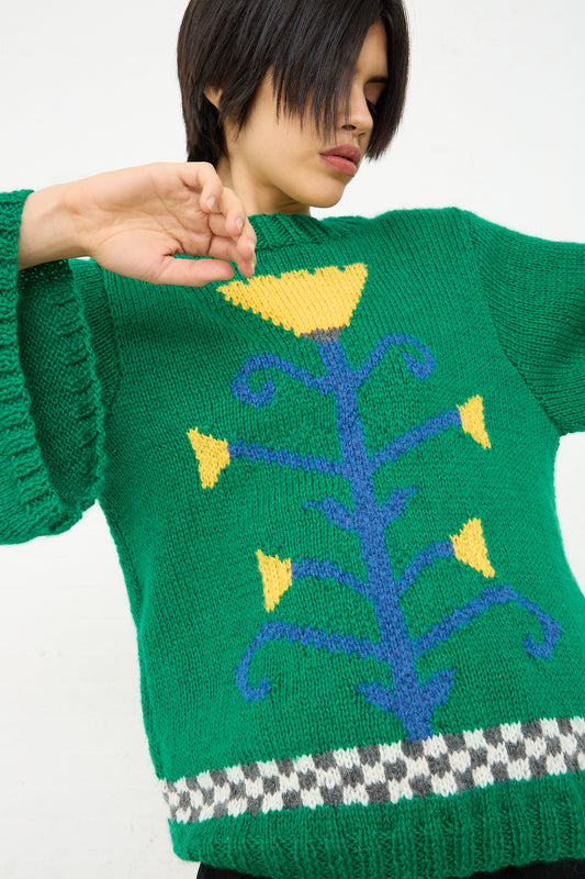 A person in a Sofio Gongli hand-knit Green Flower Sweater with a yellow and blue floral design pulling at the collar.