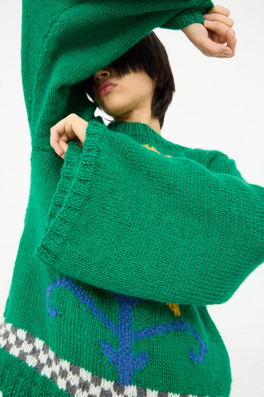 A person in a Sofio Gongli Yellow Flower Sweater in Green with embroidered details is lifting the sweater over their head, partially obscuring their face. Only the lower half of the person's face is visible.