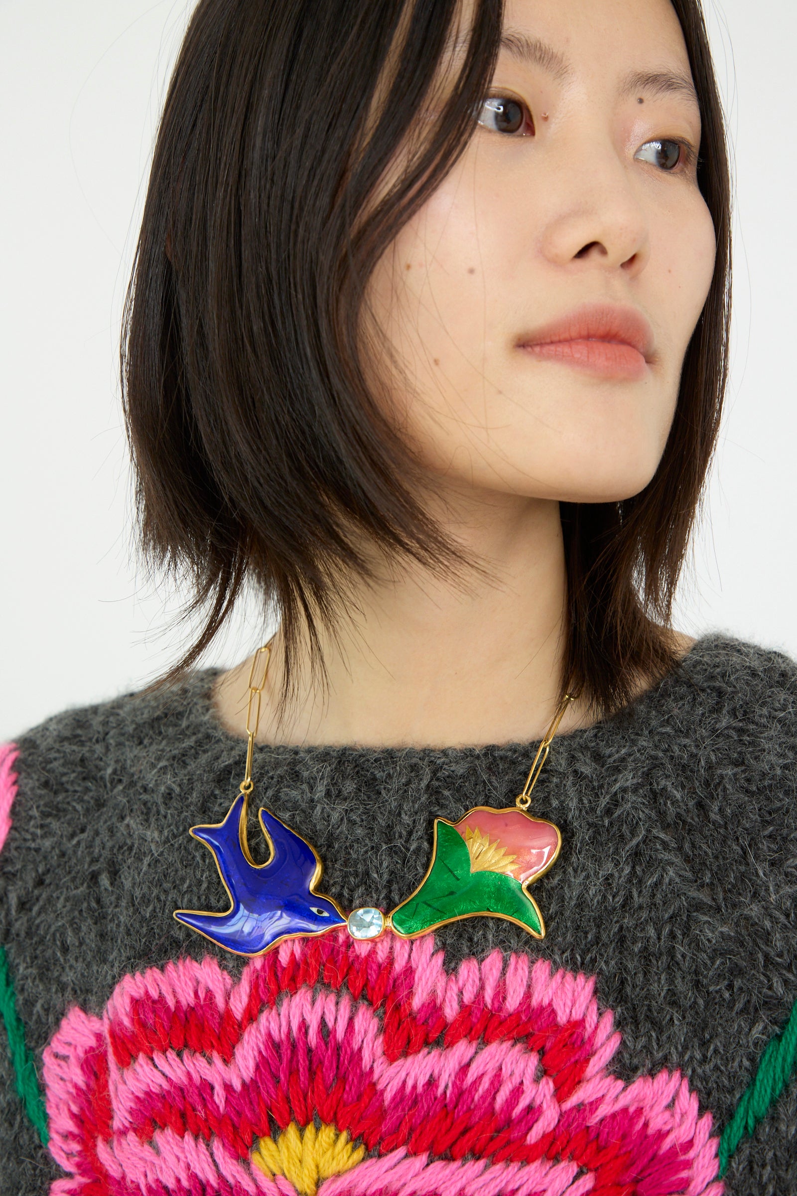 A person with short hair wears a gray sweater embellished with pink flowers, complemented by the Sofio Gongli Bird and Flower Necklace.