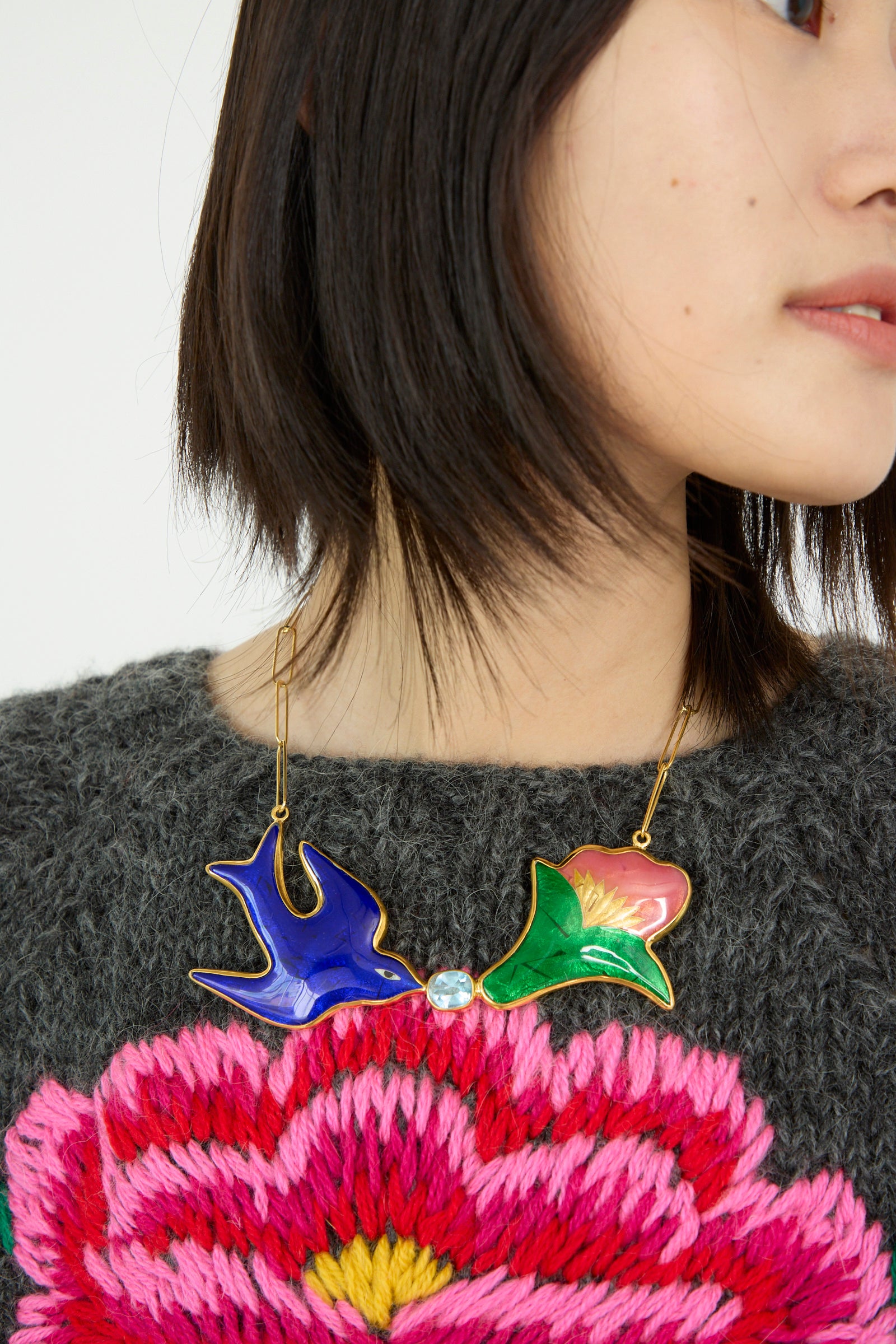 A person wearing a vibrant Sofio Gongli Bird and Flower Necklace paired with a sweater adorned with a large pink flower design.