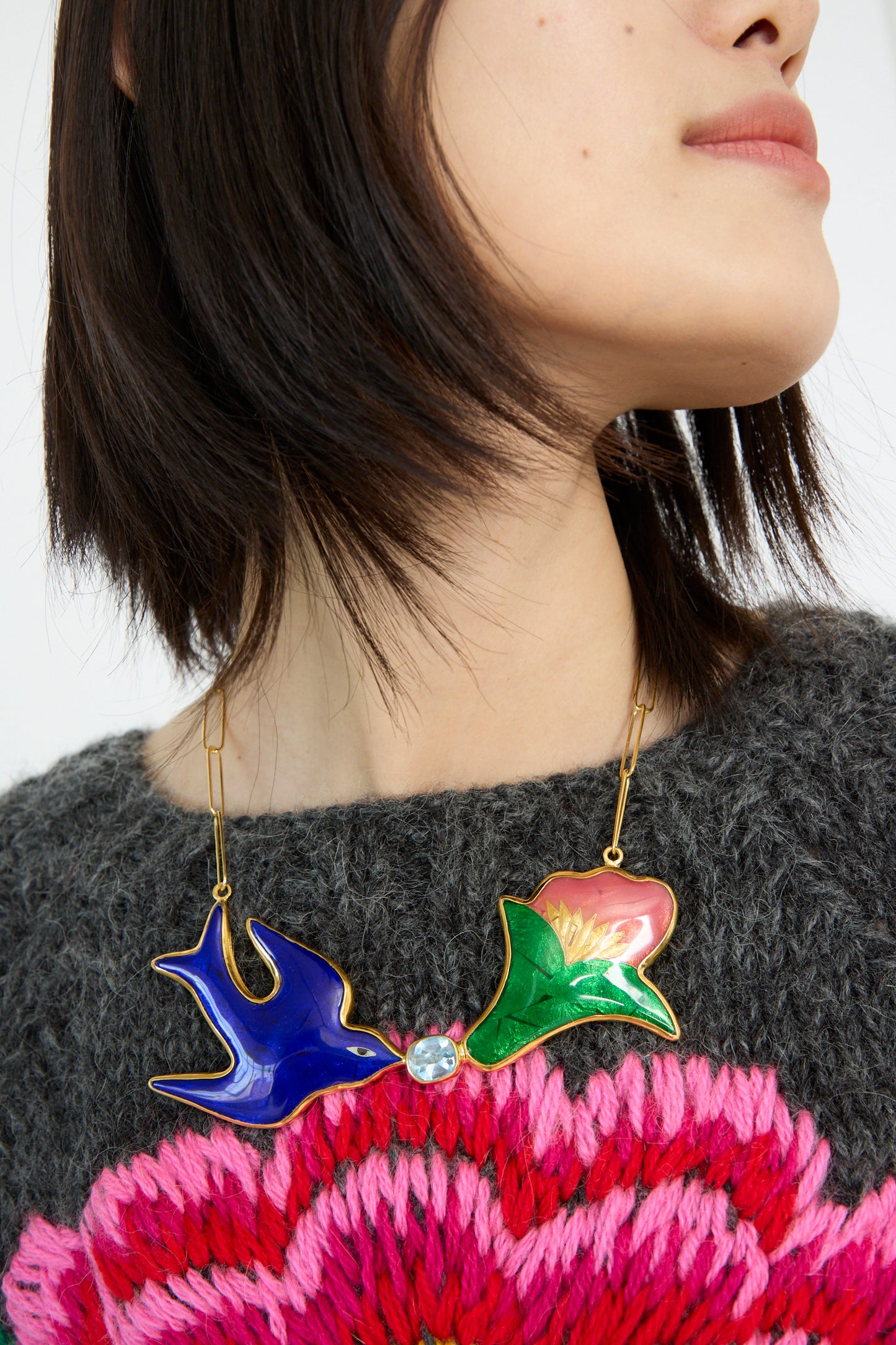 Someone wears a Sofio Gongli Bird and Flower Necklace, featuring vibrant birds and flowers, paired with a gray and pink knitted sweater.