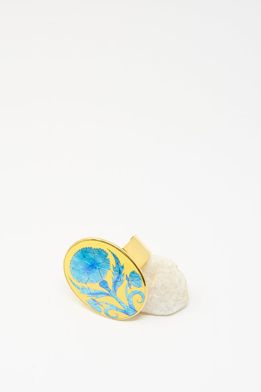 The Sofio Gongli Flower Bracelet in Blue is inspired by Byzantine cloisonné art, featuring a blue floral design, and is displayed upright on a white stone with a clean white background.