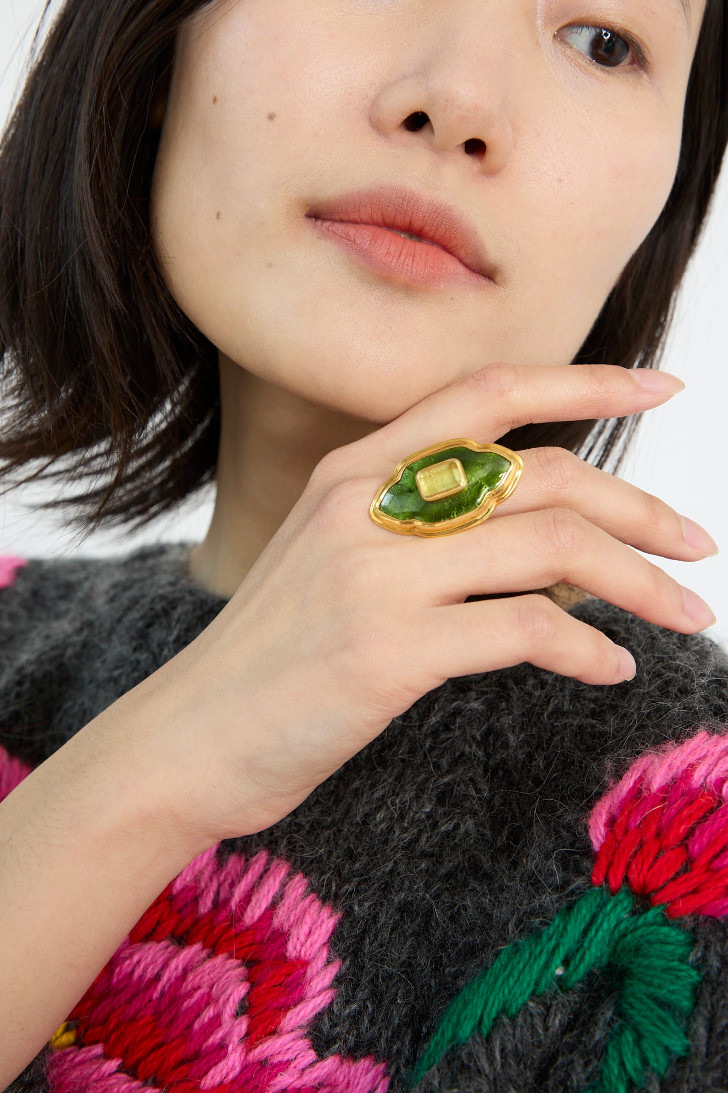 A person with a Sofio Gongli Ring in Green and Gold holds a hand near their face while wearing a grey sweater with pink and red floral patterns.