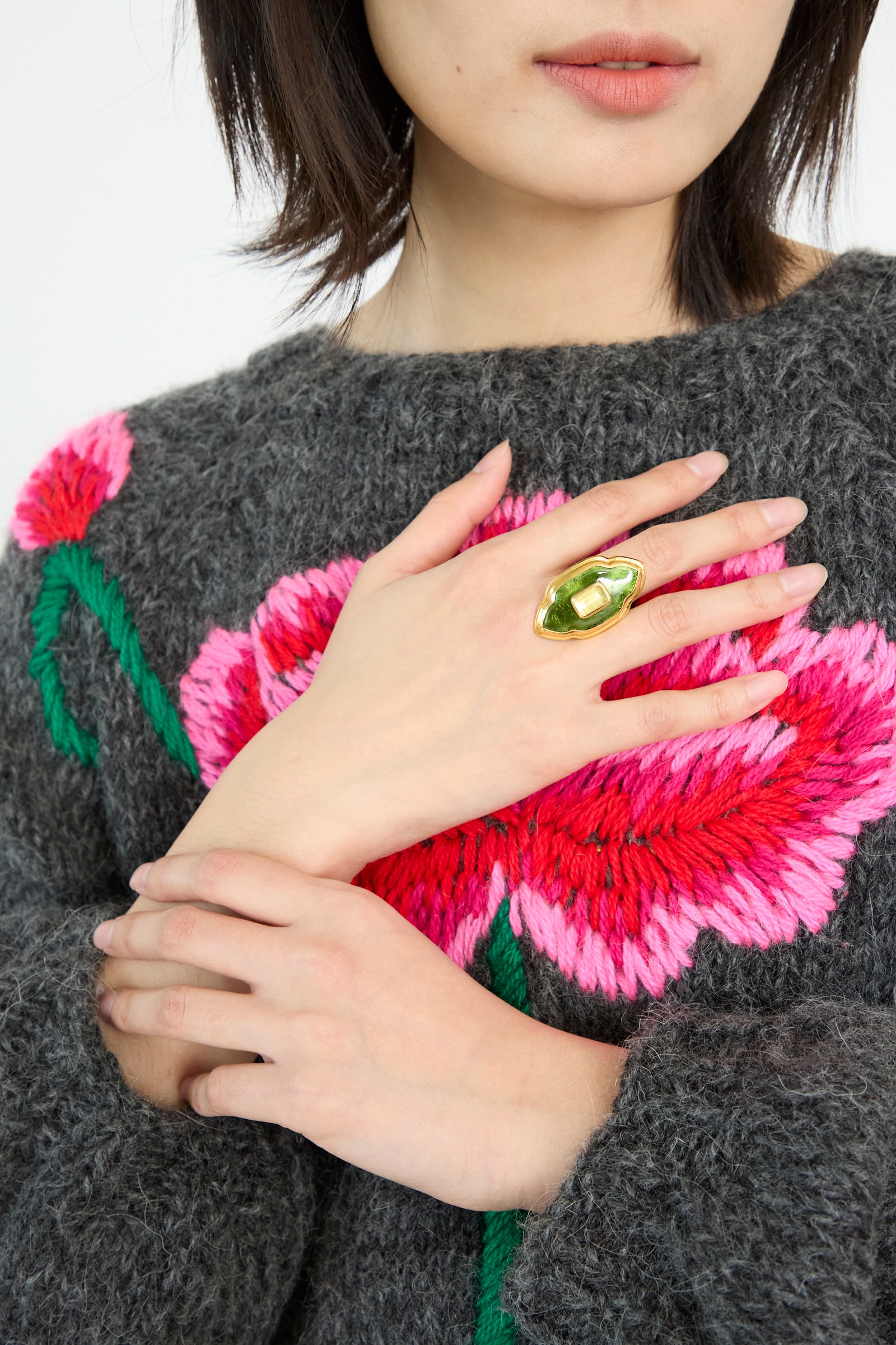 A model wears a sweater with pink floral patterns, while Sofio Gongli's Ring in Green and Gold sits on their middle finger, hand on their chest against a white backdrop. 