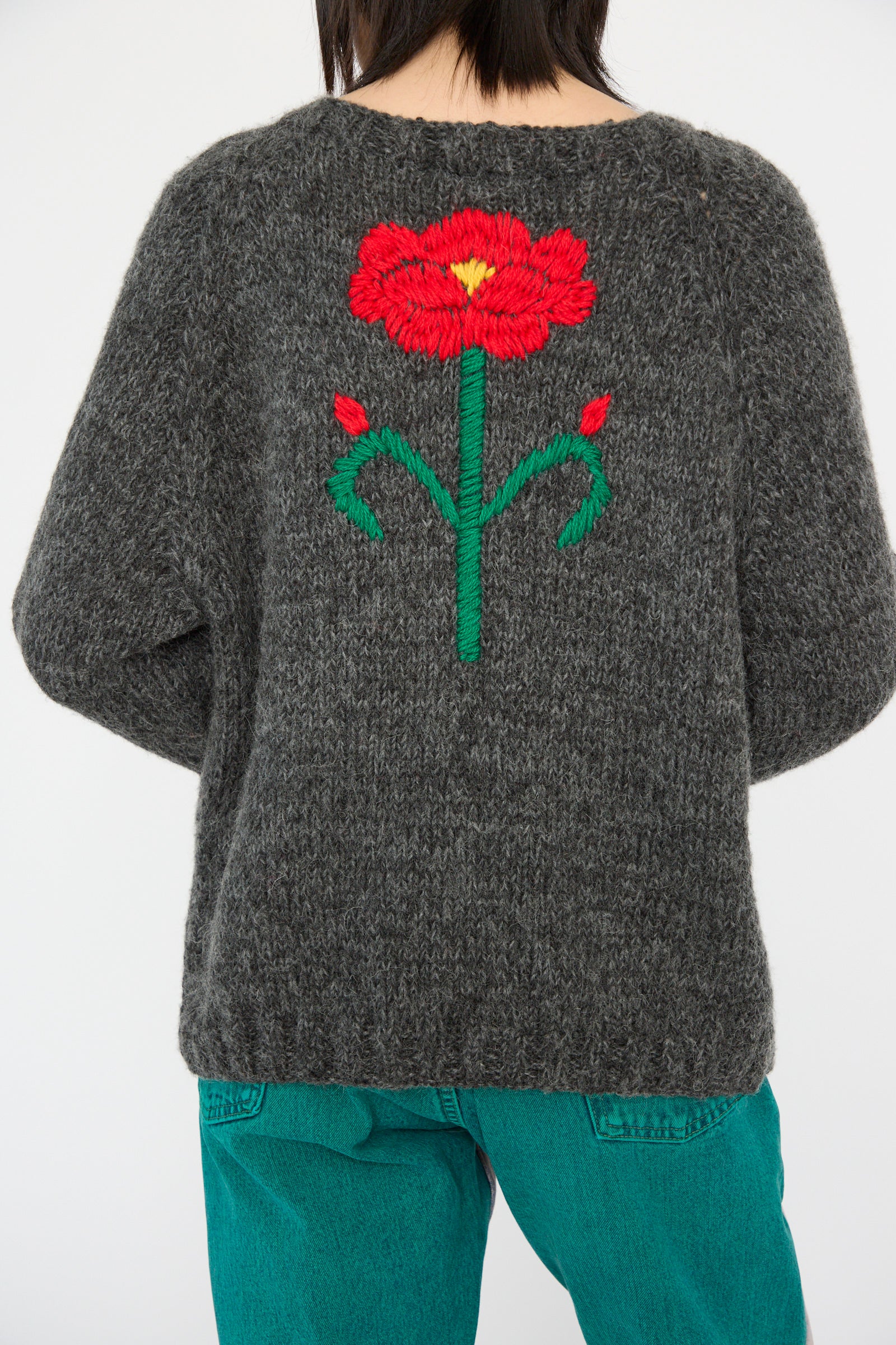 A person is wearing the Sofio Gongli Sweater with Red and Pink Flower in Charcoal, paired with teal pants.