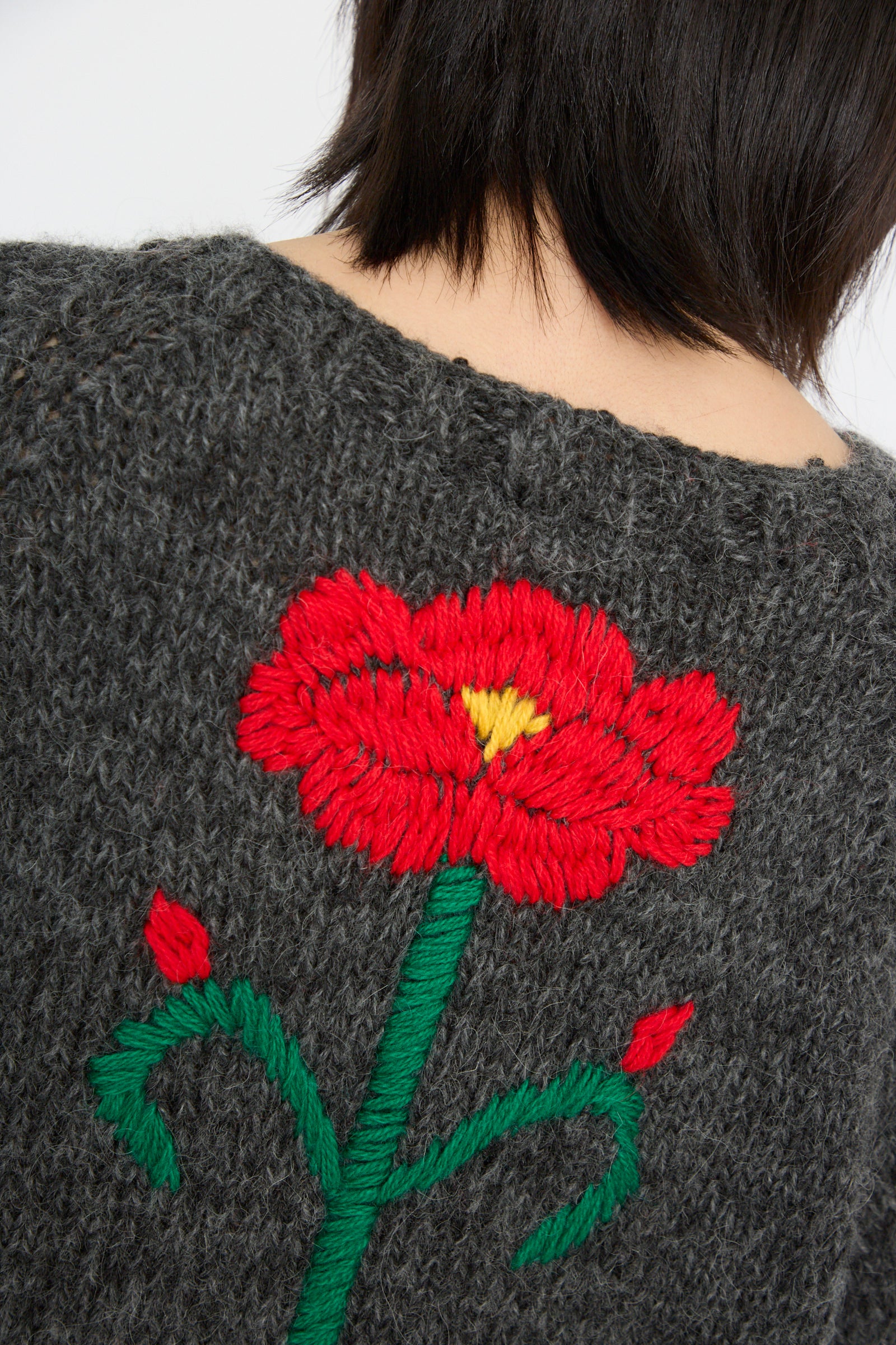 Close-up of a person wearing the Sofio Gongli "Sweater with Red and Pink Flower in Charcoal," featuring a hand-knit mohair wool blend design with an exquisite embroidered red flower and green stem.