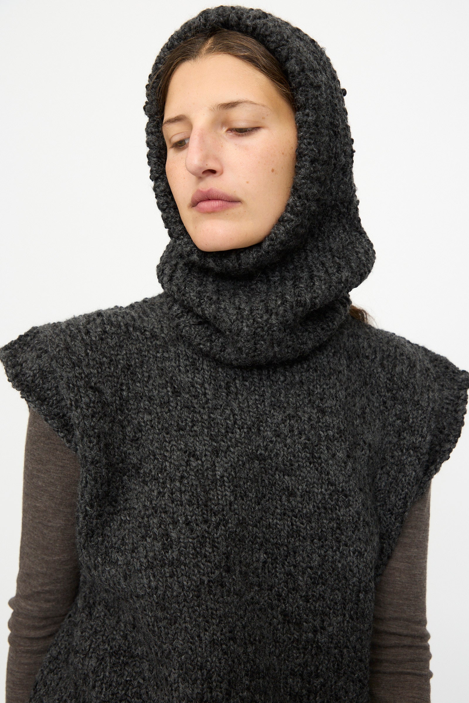 A woman models the SONO Alpaca and Wool Bruna Balaclava in Charcoal, wearing it over a long-sleeve brown shirt and looking down with a neutral expression against a plain background.
