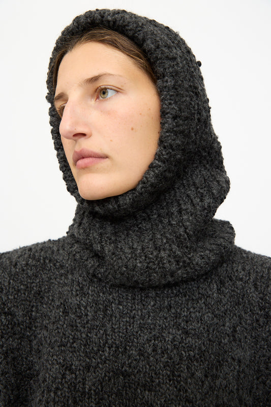 A person wearing the SONO Alpaca and Wool Bruna Balaclava in Charcoal, a chunky knit hood crafted from an alpaca-wool blend, looks slightly to the side against a plain background.