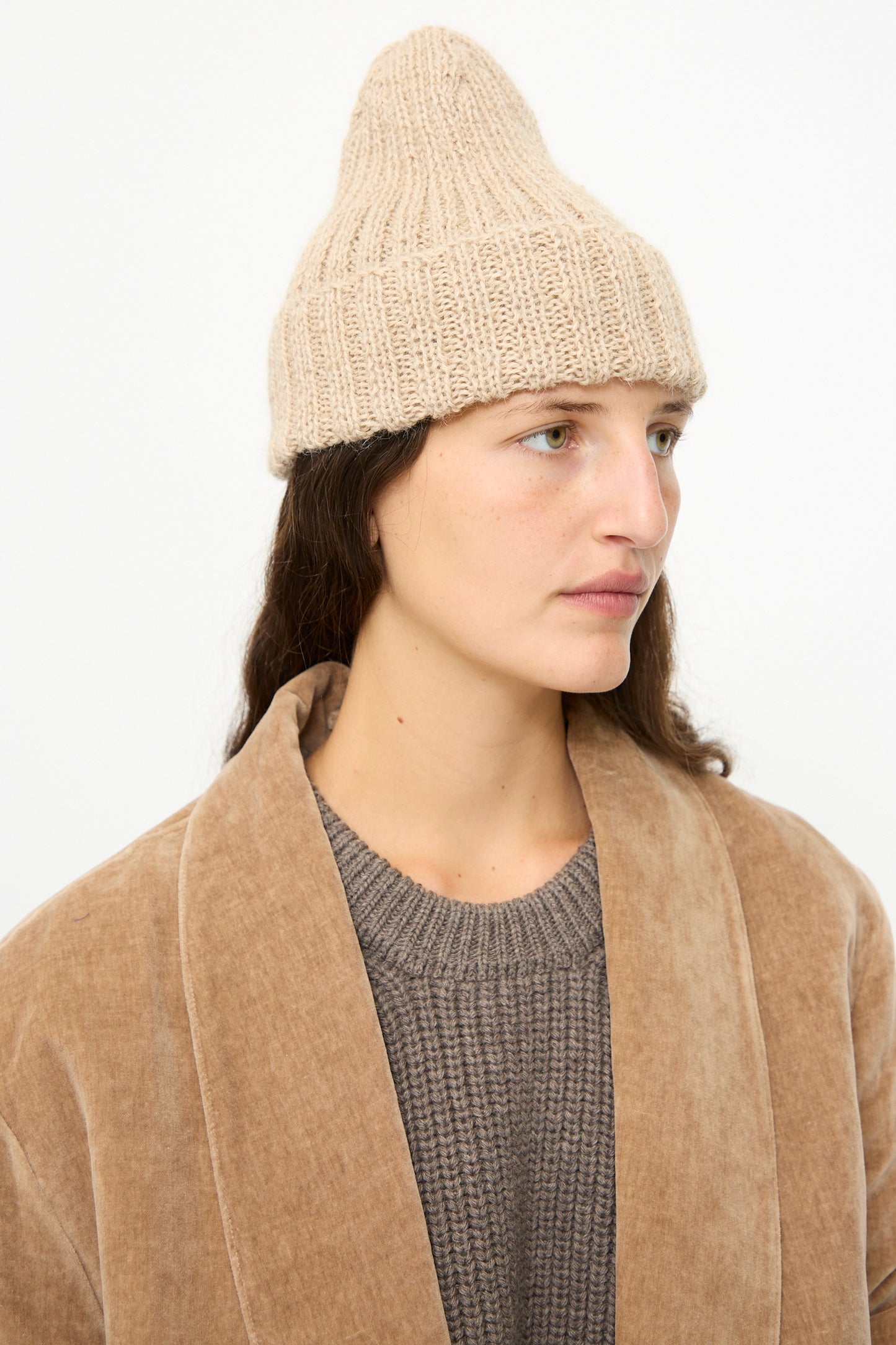 A person wearing the SONO Alpaca and Wool Gyu Beanie in Oat, a brown jacket, and a gray sweater stands against a plain white background, looking to the side.