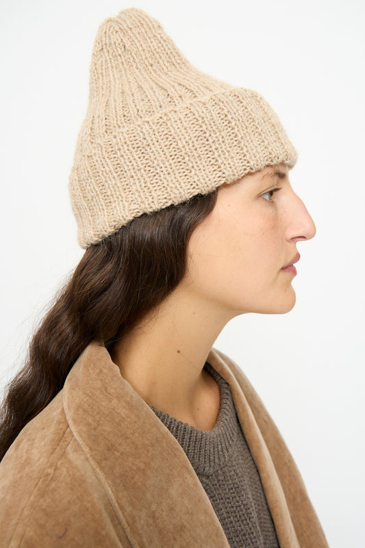 A person with long brown hair wears the SONO Alpaca and Wool Gyu Beanie in Oat, a brown sweater, and a tan coat, shown in profile against a plain light background.