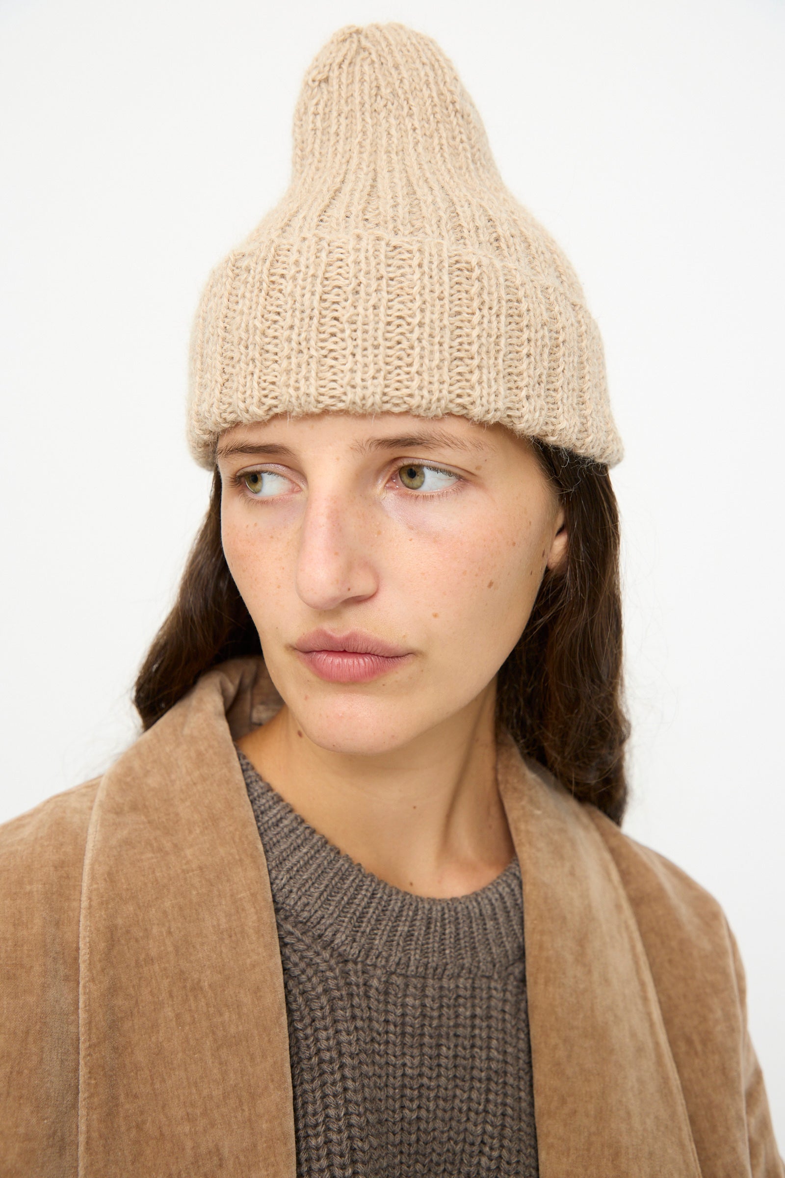 A person wearing the SONO Alpaca and Wool Gyu Beanie in Oat, a brown jacket, and a gray sweater looks off to the side against a plain background.