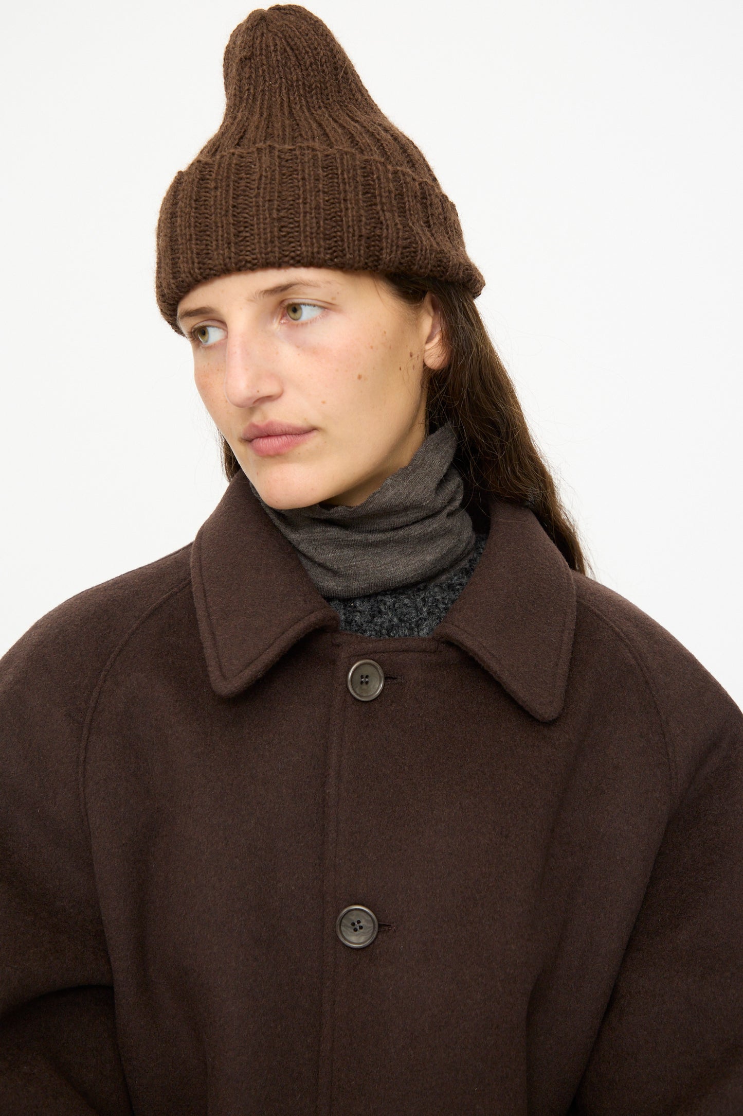A person wears the SONO Alpaca and Wool Gyu Beanie in Terra, a brown wool coat, and a dark scarf, looking to the side against a plain white background.
