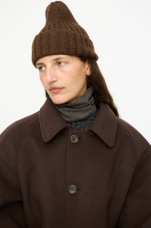 A person wears the SONO Alpaca and Wool Gyu Beanie in Terra, a brown wool coat, and a dark scarf, looking to the side against a plain white background.