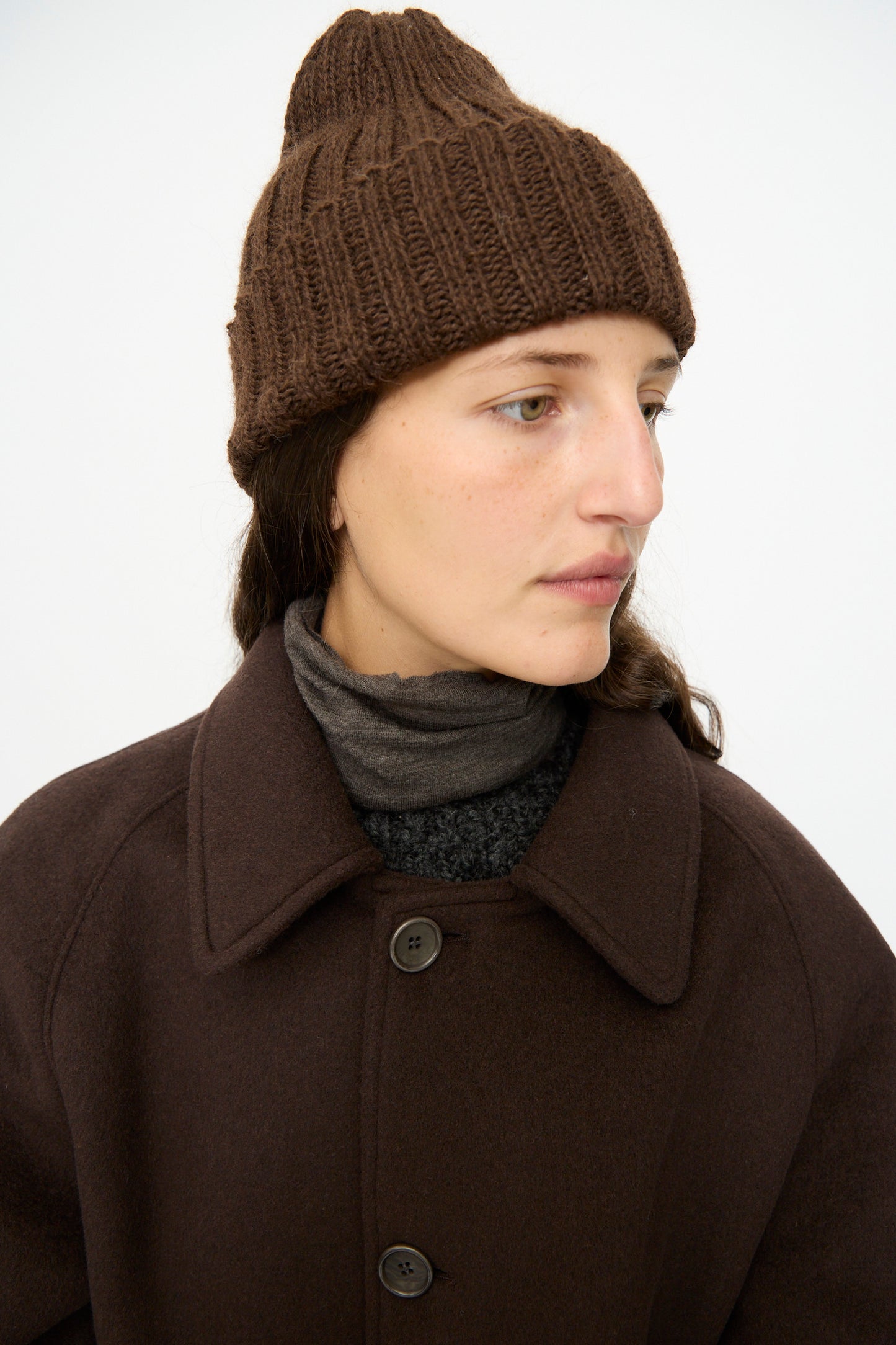 A person wearing the SONO Alpaca and Wool Gyu Beanie in Terra, a dark brown coat, and layered turtlenecks looks off to the side against a plain background.