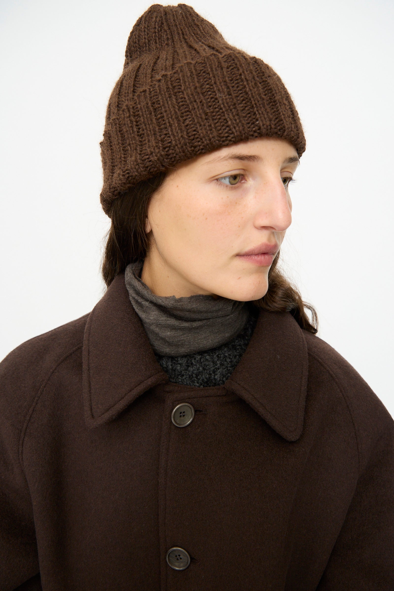 A person wearing the SONO Alpaca and Wool Gyu Beanie in Terra, a dark brown coat, and layered turtlenecks looks off to the side against a plain background.