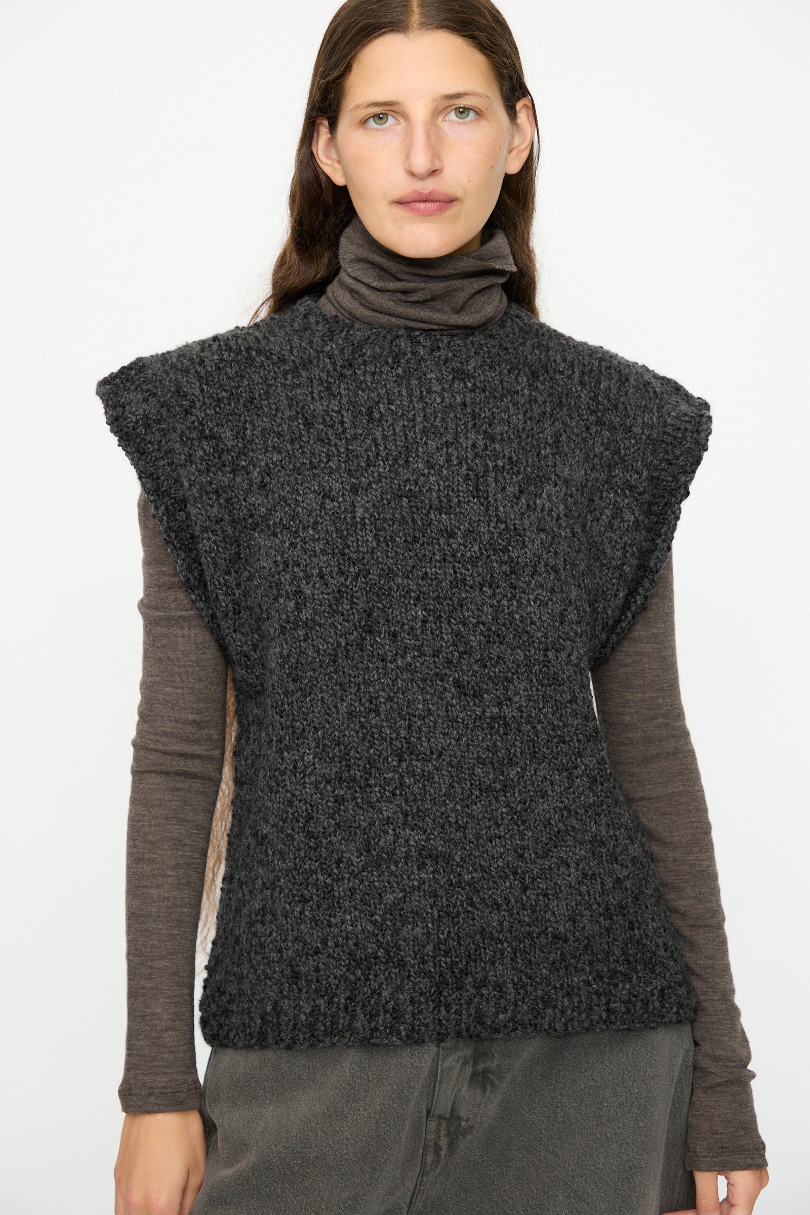 A person with long brown hair wears the SONO Alpaca and Wool Petras Pullunder in Charcoal, made in the UK, layered over a brown turtleneck and gray pants, standing against a plain white background.