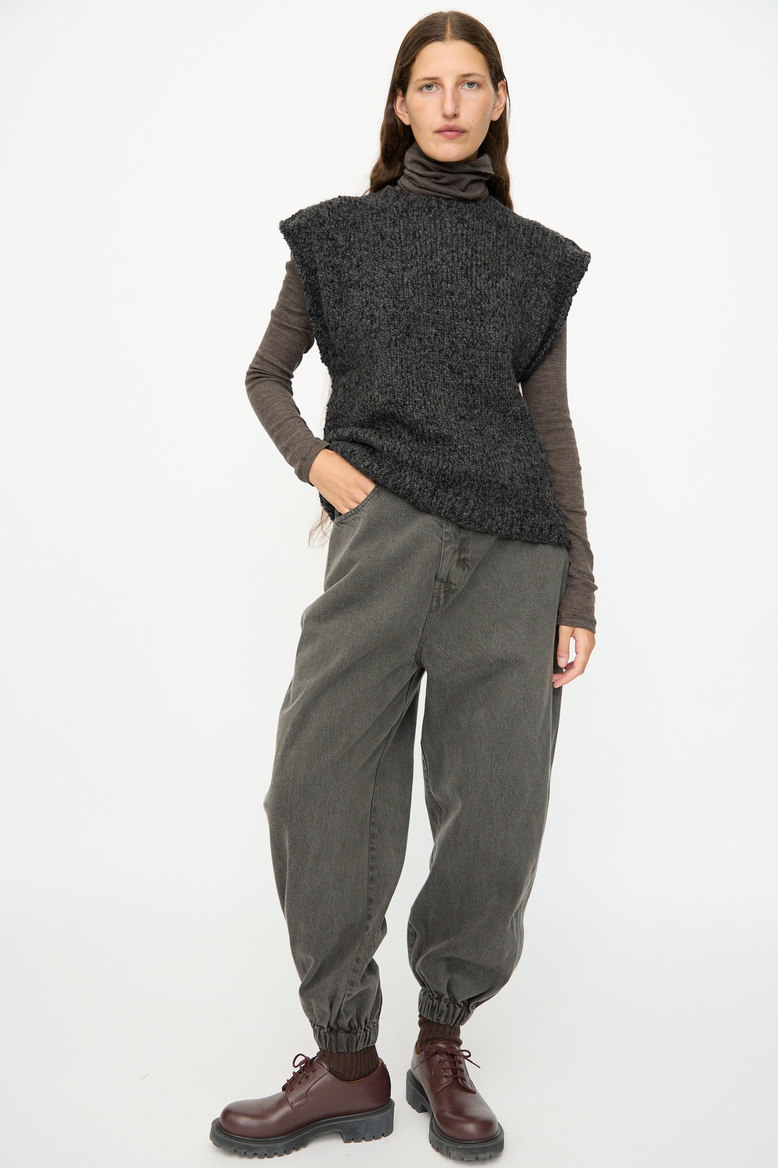 A person stands against a plain background wearing the SONO Alpaca and Wool Petras Pullunder in Charcoal over a brown turtleneck, paired with loose gray pants with elasticated cuffs and brown lace-up boots.