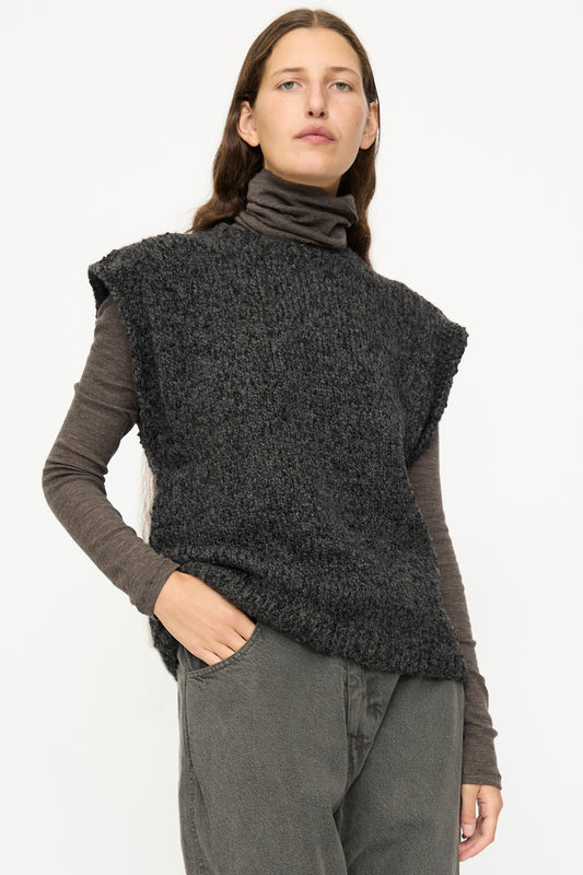 A person with long brown hair wears the SONO Alpaca and Wool Petras Pullunder in Charcoal over a brown turtleneck and gray pants, standing against a plain white background.