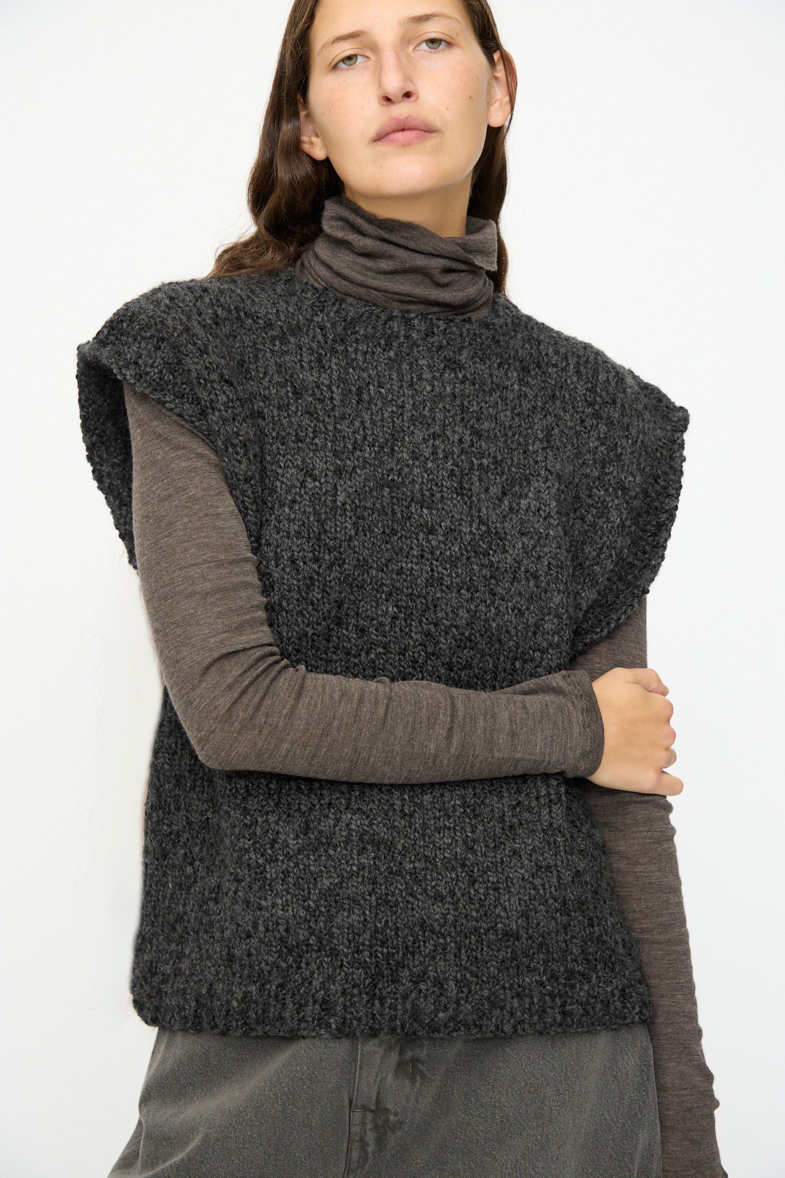 A person wears the SONO Alpaca and Wool Petras Pullunder in Charcoal over a long-sleeved brown turtleneck, standing against a plain white background.