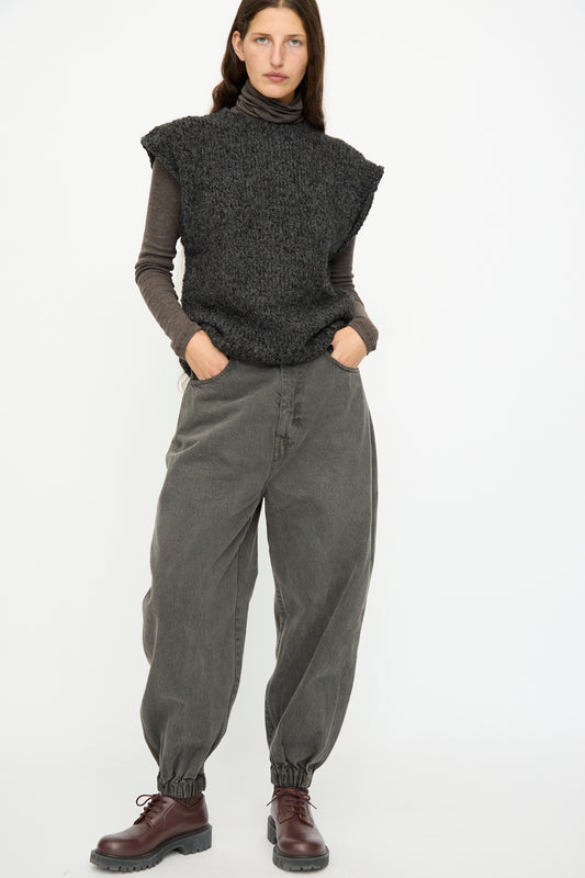 A woman stands against a white background wearing SONO's Cotton Jah Jeans in Off Black—baggy, elastic-waist jeans with cinched ankles—paired with a dark sleeveless sweater over a turtleneck and brown lace-up boots.