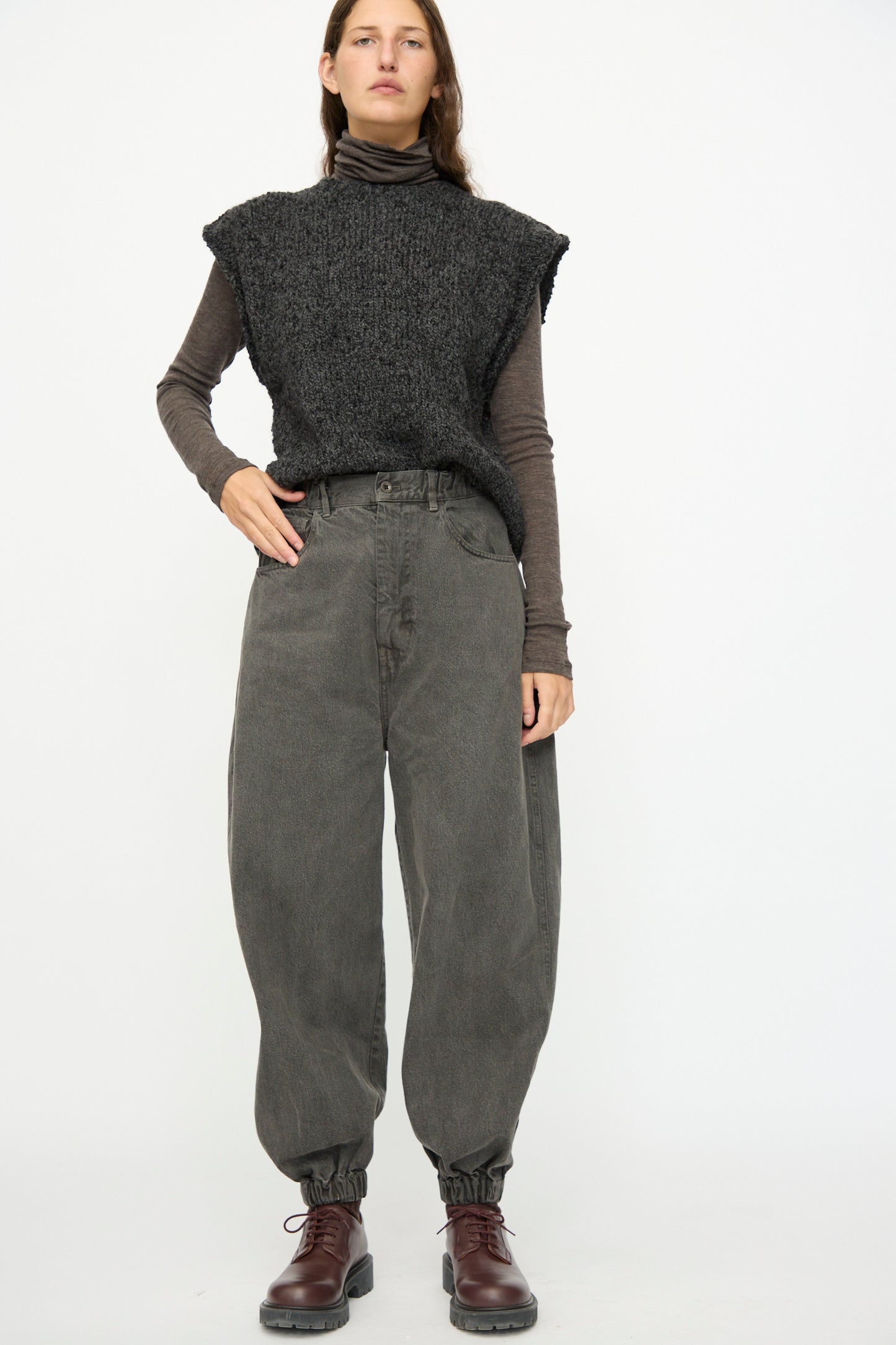 A person wearing SONO's Cotton Jah Jeans in Off Black, paired with a dark knit vest, brown long-sleeve shirt, and brown lace-up shoes stands against a plain white background.