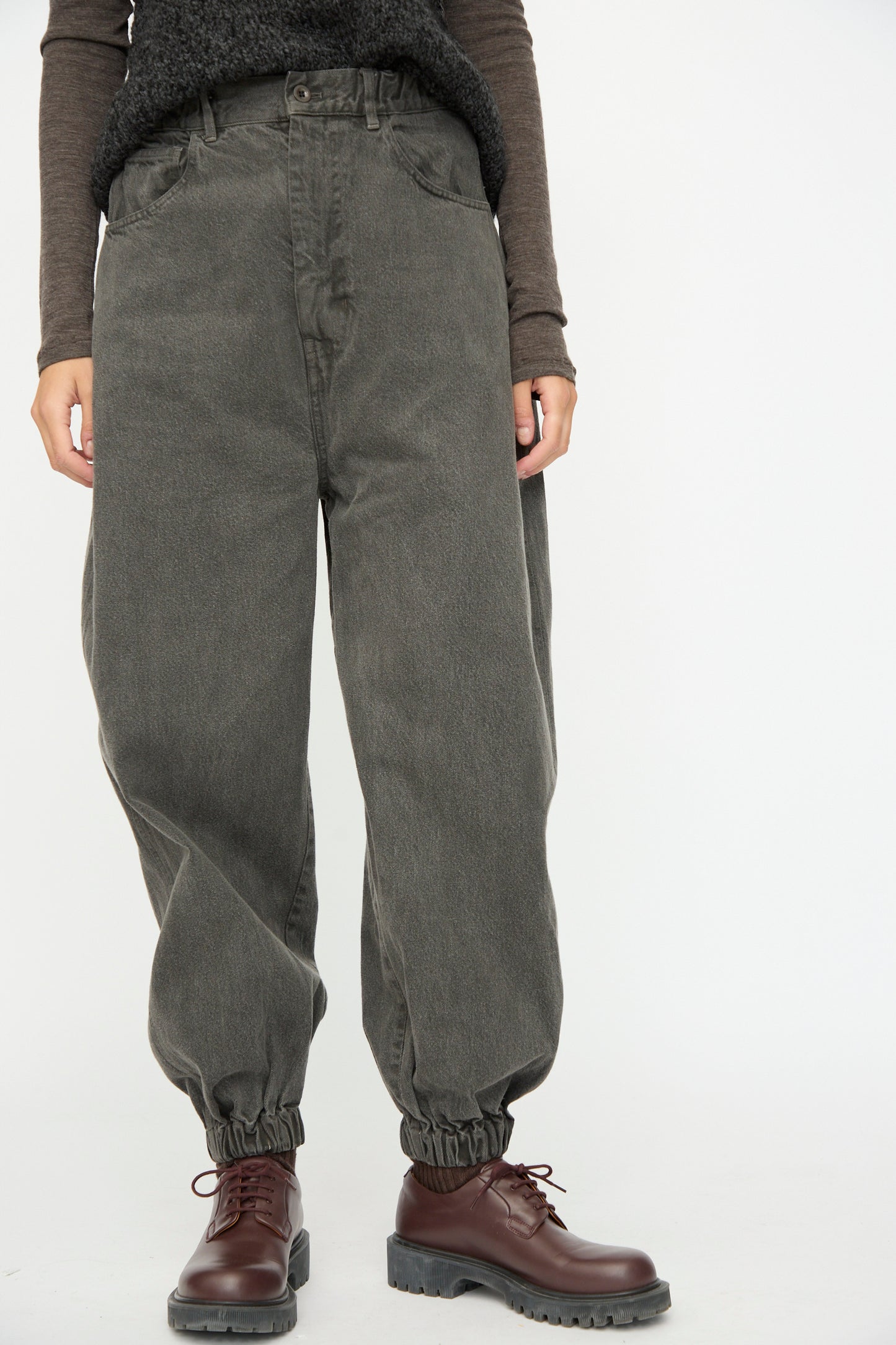 A person wears SONO's Cotton Jah Jeans in Off Black with a brown long-sleeve top and brown lace-up boots, standing against a plain white background.