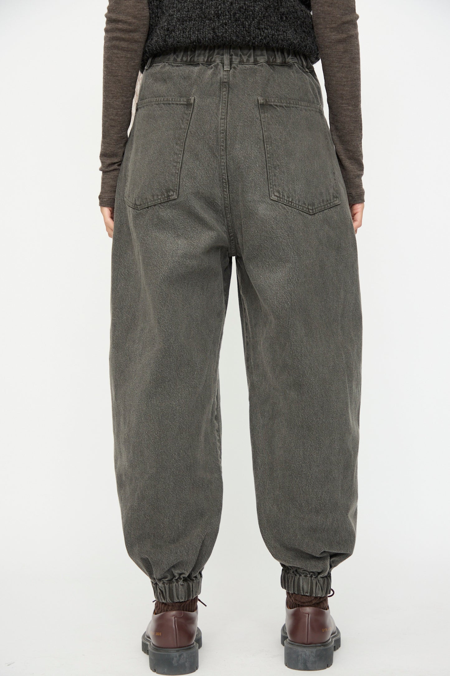 A person stands facing away against a plain white background, wearing SONO Cotton Jah Jeans in Off Black with an elasticated waist, paired with brown shoes and a long-sleeve brown top.