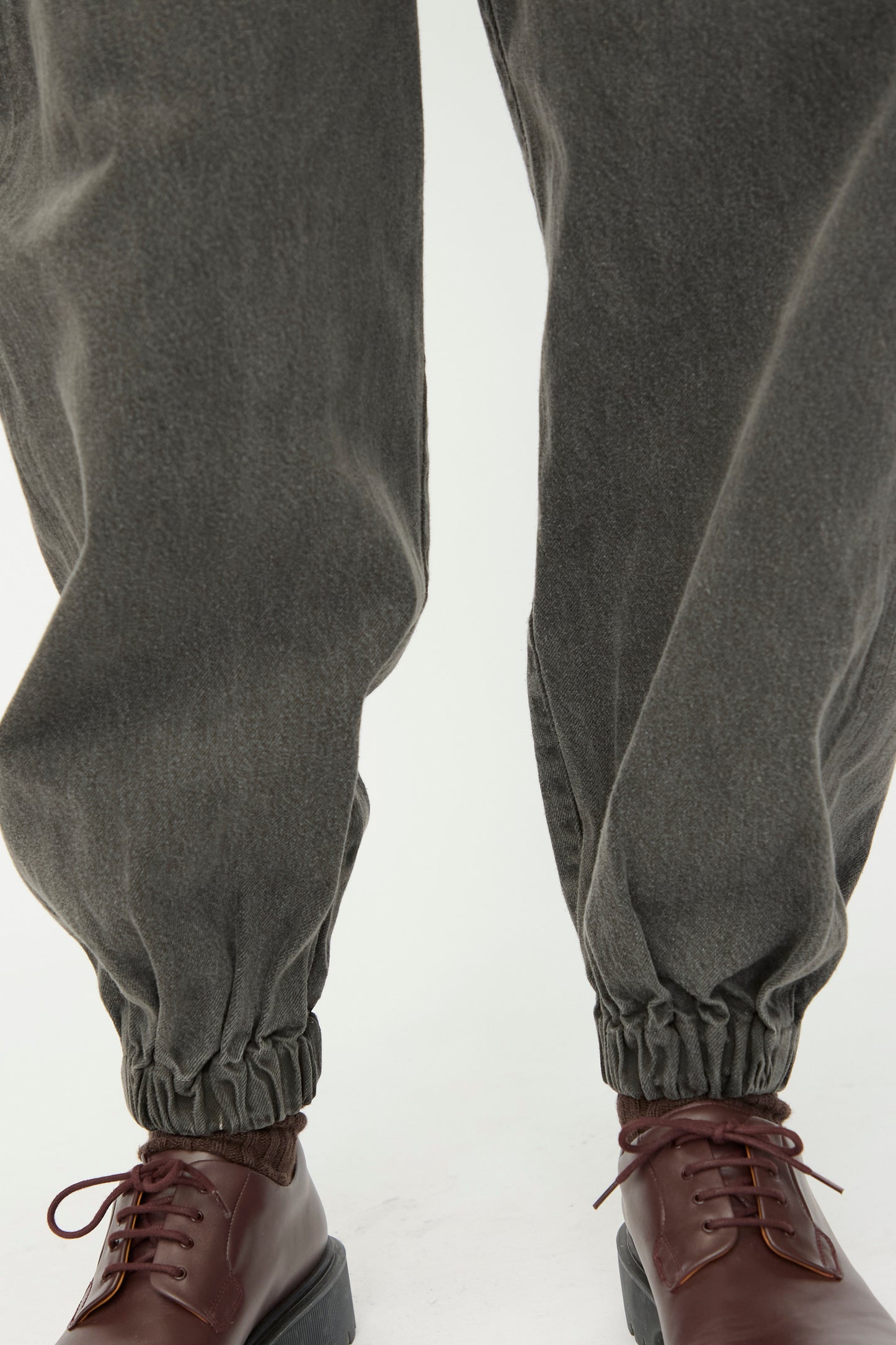 Close-up of a person wearing SONO’s Cotton Jah Jeans in Off Black with an elasticated waist and cuffs, paired with brown lace-up shoes, standing on a light-colored surface.