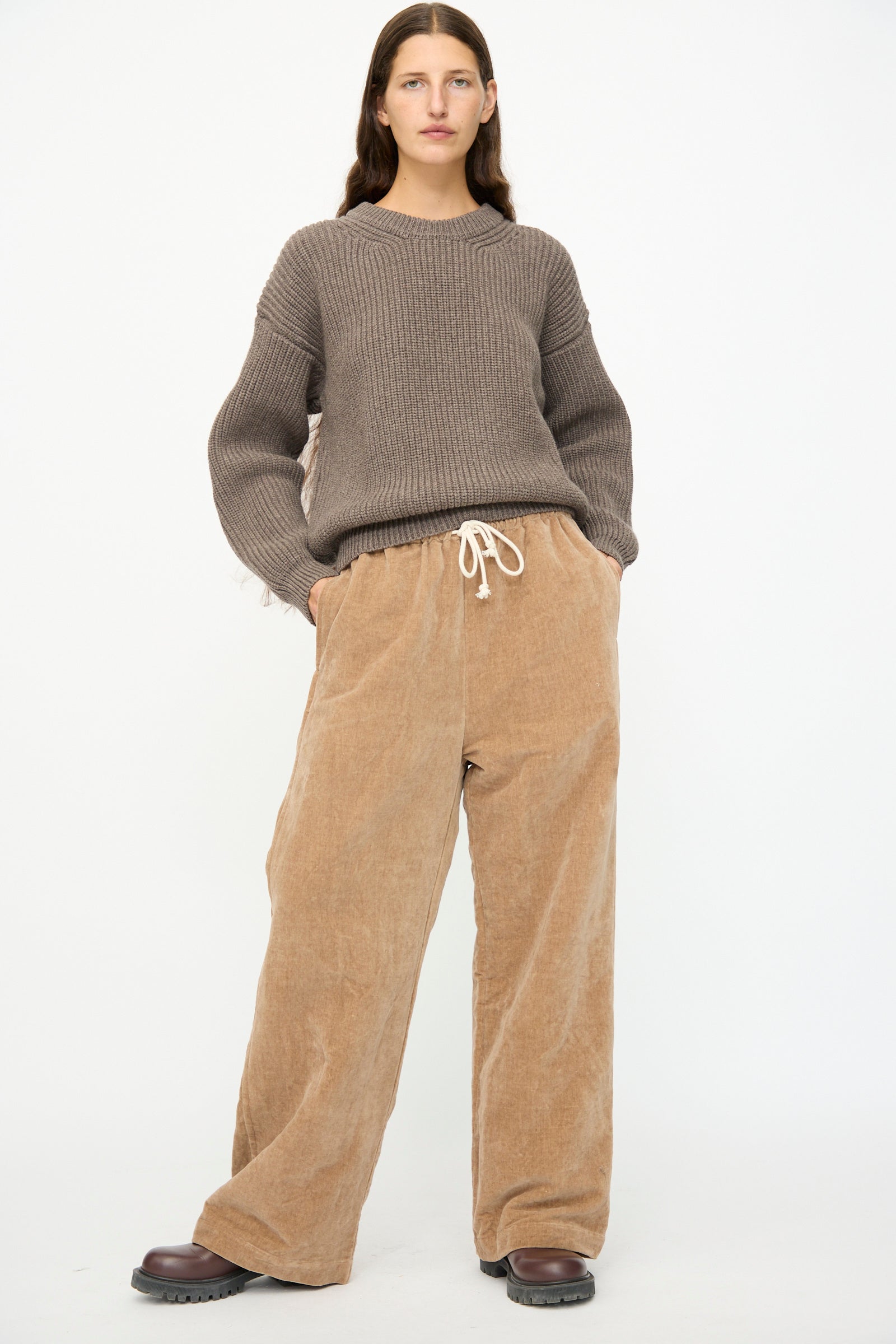 A person stands against a white background wearing SONO's Johnny Jogger in Desert Rose—relaxed fit corduroy pants with a drawstring waist—paired with a brown knit sweater and dark brown shoes.