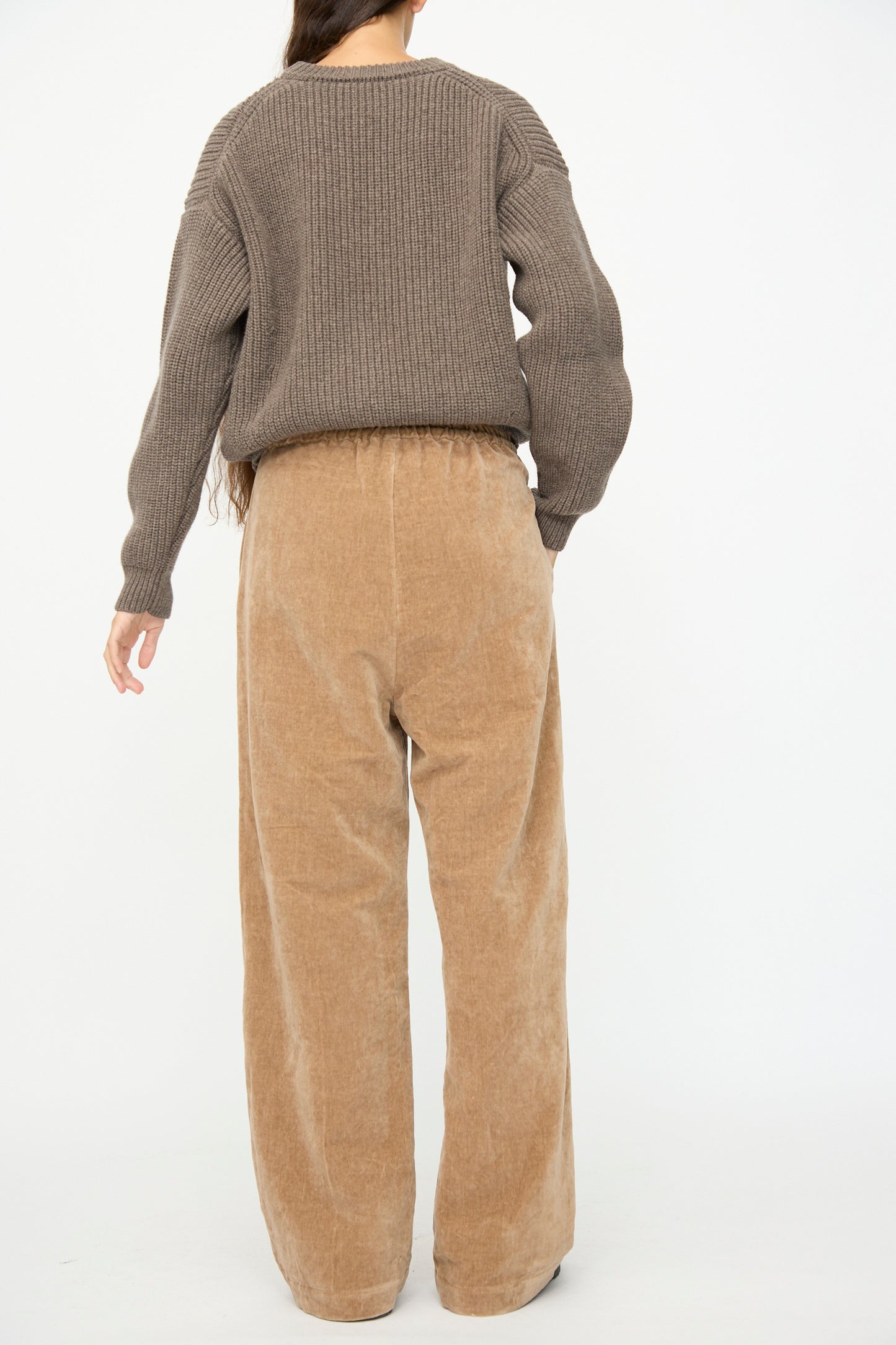 A person stands with their back to the camera against a white background, wearing SONO's Johnny Jogger in Desert Rose—relaxed fit pants with a drawstring waist—and a brown ribbed sweater.