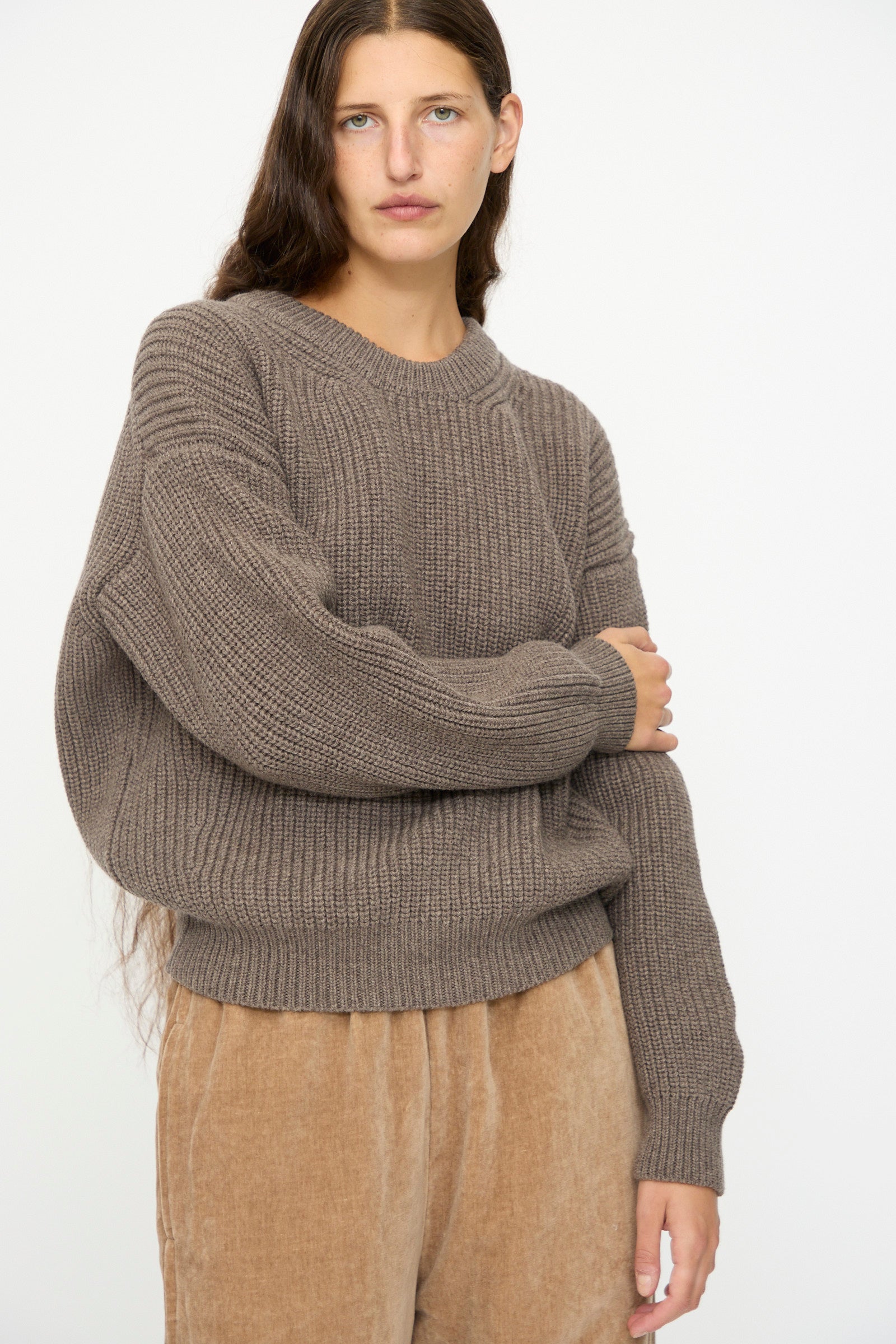 A person with long brown hair wears the SONO Natural Wool Joel Jumper in Smoke, featuring a chunky ribbed design and oversized fit, paired with light brown corduroy pants against a plain white background.