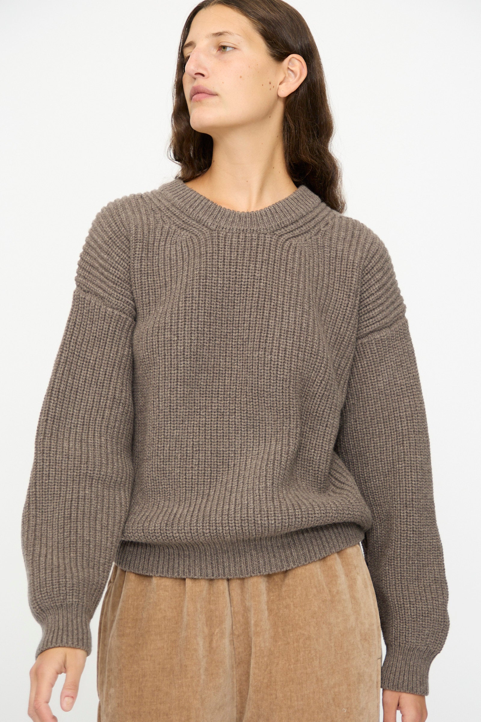 A person with long brown hair models the SONO Natural Wool Joel Jumper in Smoke, paired with tan corduroy pants, against a plain light background.