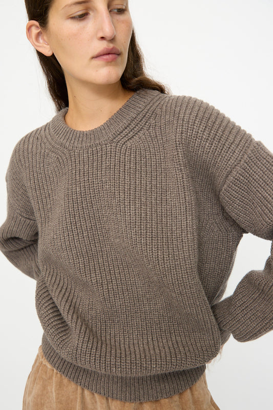 A woman wears the SONO Natural Wool Joel Jumper in Smoke with light brown pants, standing against a plain white background.