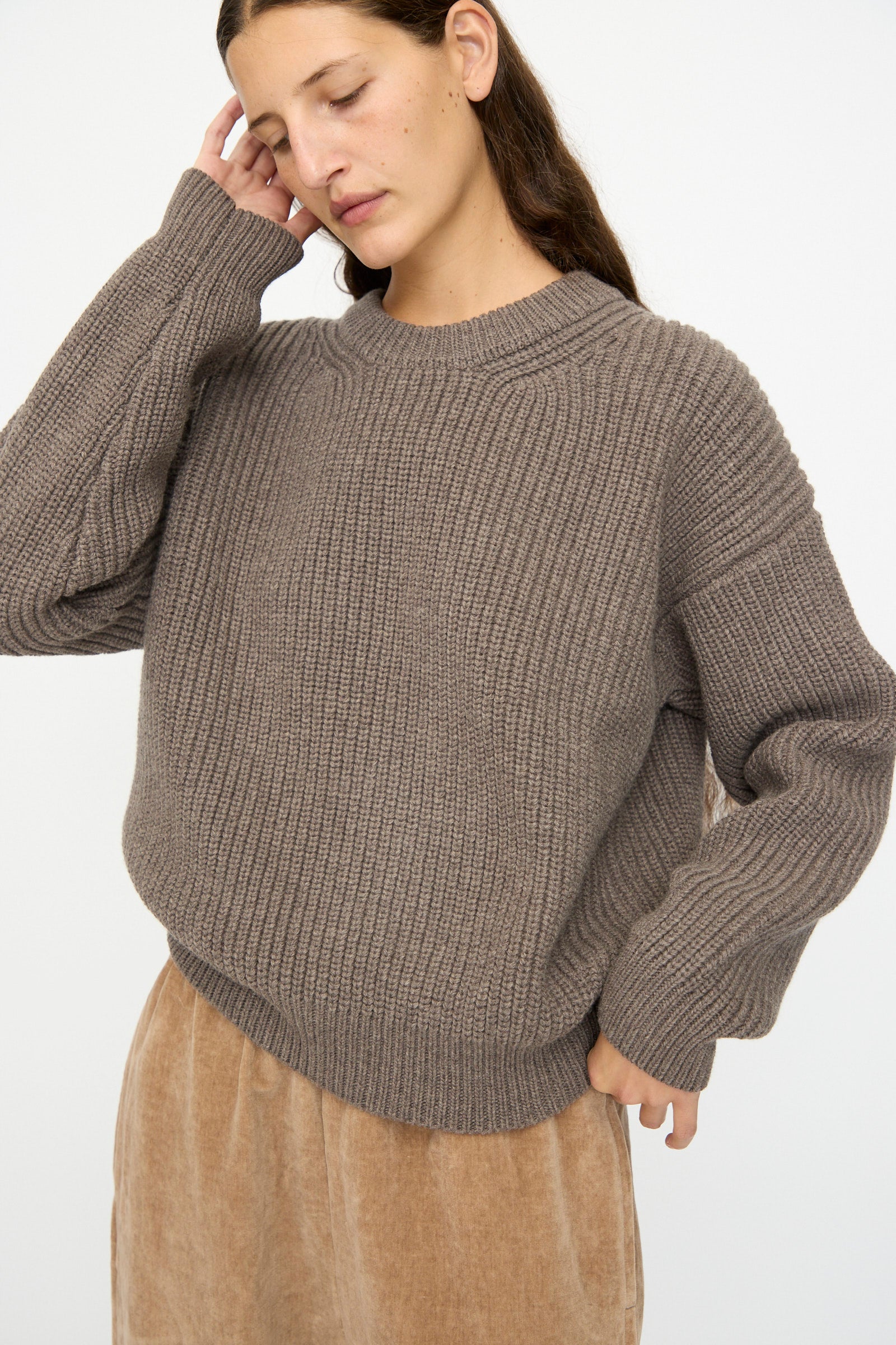 A woman with long brown hair wears the SONO Natural Wool Joel Jumper in Smoke, styled oversized, with a beige skirt, standing against a plain white background.