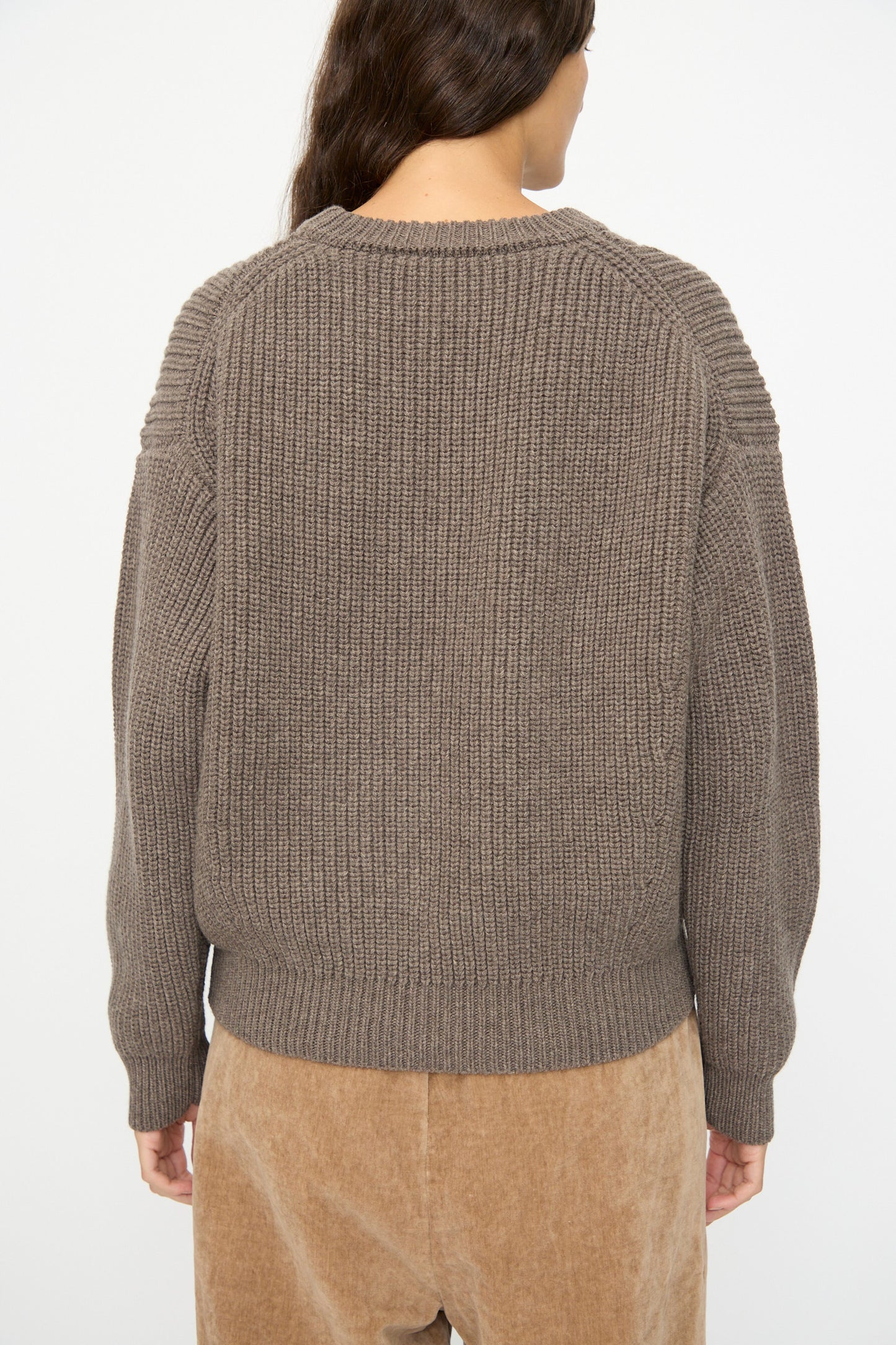 A person with long brown hair wears the SONO Natural Wool Joel Jumper in Smoke, styled oversized, paired with tan corduroy pants, photographed from the back against a plain white background.