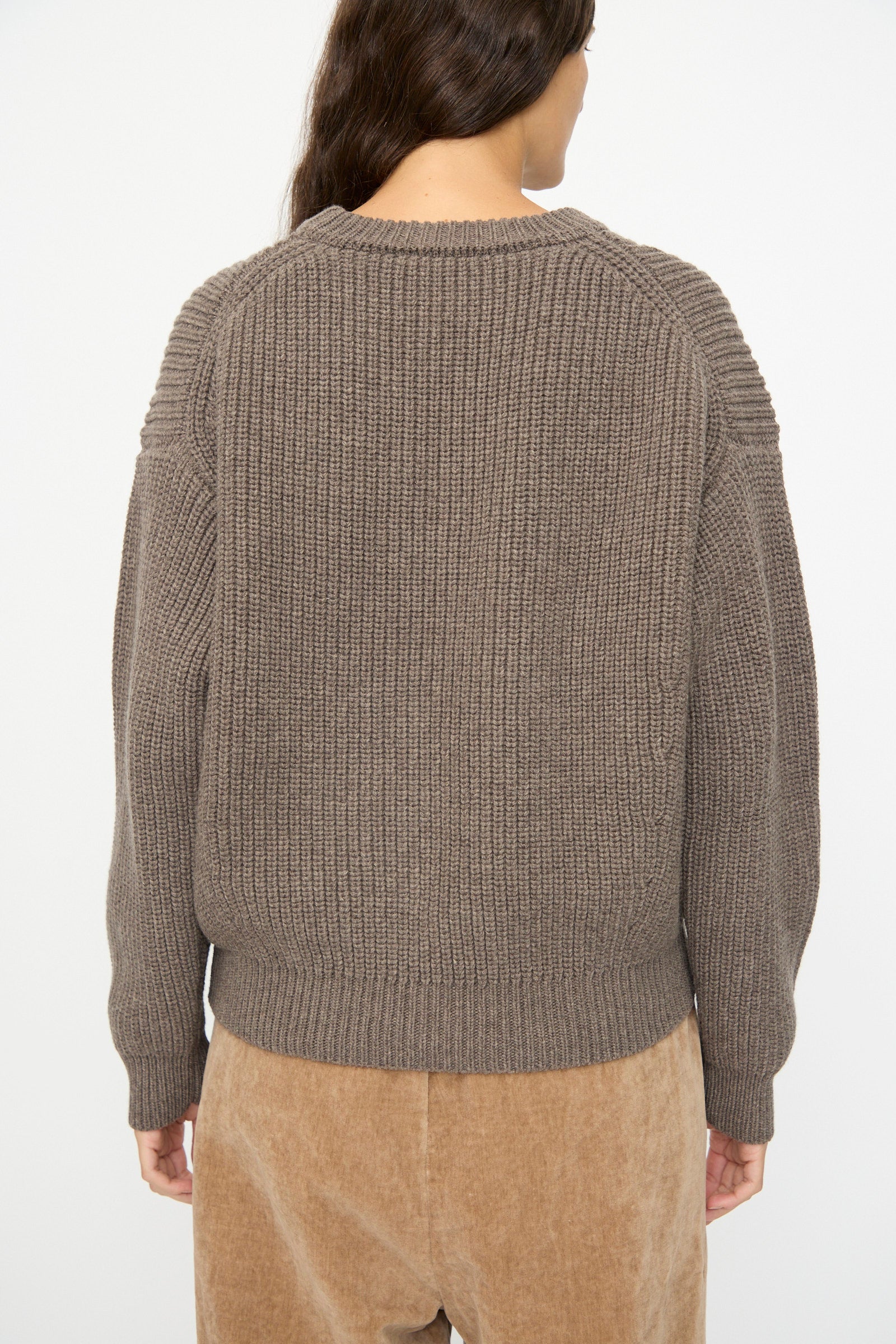 A person with long brown hair wears the SONO Natural Wool Joel Jumper in Smoke, styled oversized, paired with tan corduroy pants, photographed from the back against a plain white background.