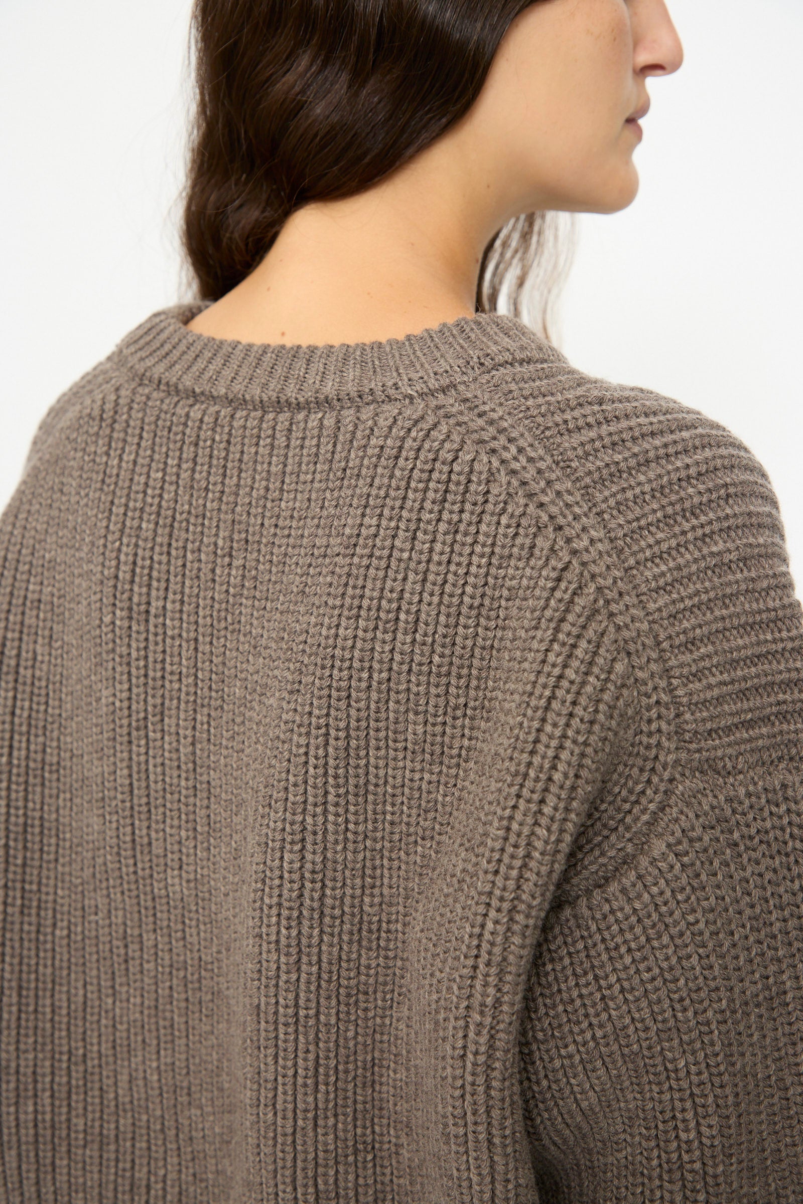 A person with long brown hair models the SONO Natural Wool Joel Jumper in Smoke, shown from the side and back against a plain background.