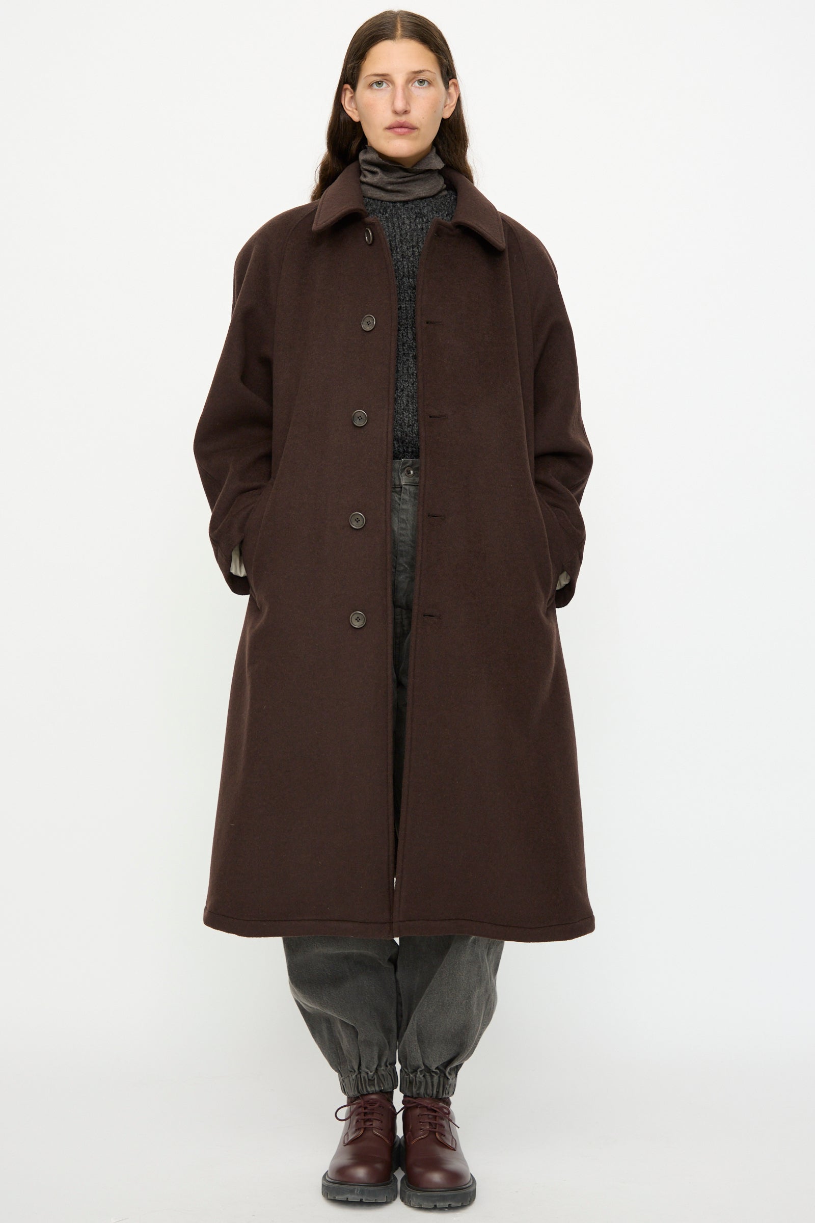 A person faces forward wearing the SONO Recycled Wool Carl Coat in Earth, paired with grey elastic-cuff pants, a dark sweater, and brown lace-up shoes in front of a plain white background.