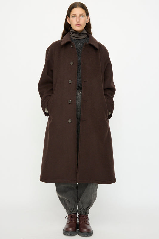 A person faces forward wearing the SONO Recycled Wool Carl Coat in Earth, paired with grey elastic-cuff pants, a dark sweater, and brown lace-up shoes in front of a plain white background.