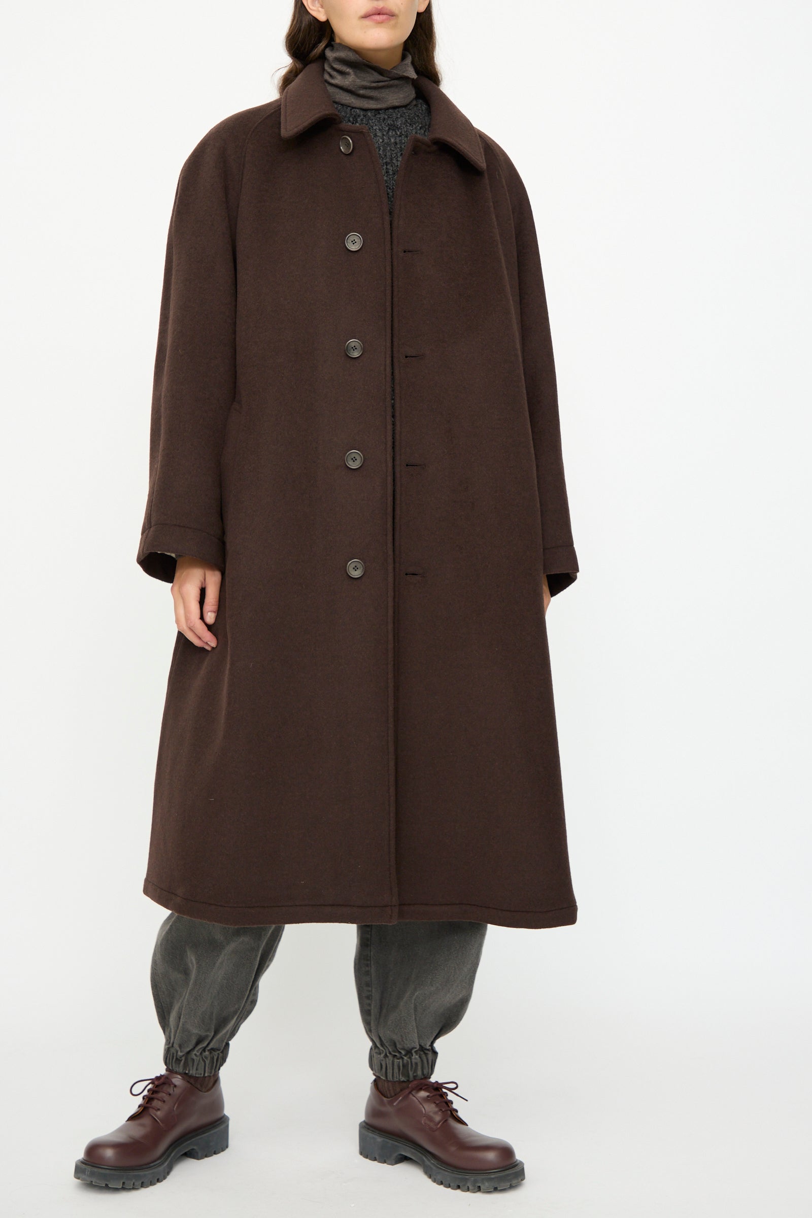 A person wears the SONO Recycled Wool Carl Coat in Earth, an oversized Italian-made piece, over a turtleneck sweater, dark elastic-cuff pants, and brown lace-up shoes, standing before a plain background.