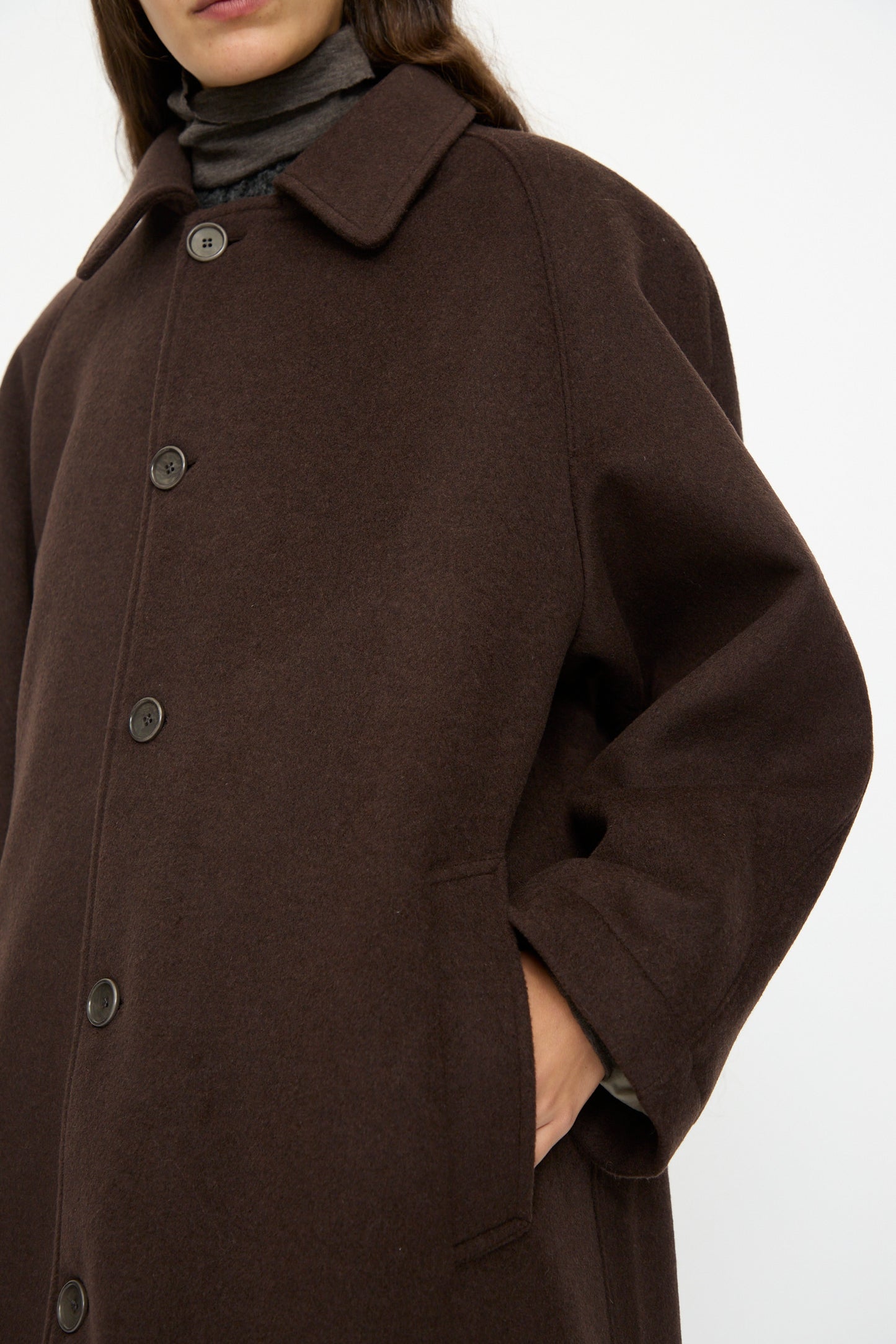 A person wearing the SONO Recycled Wool Carl Coat in Earth, an oversized dark brown coat, over a high-neck gray top with one hand in the pocket, stands against a plain background.