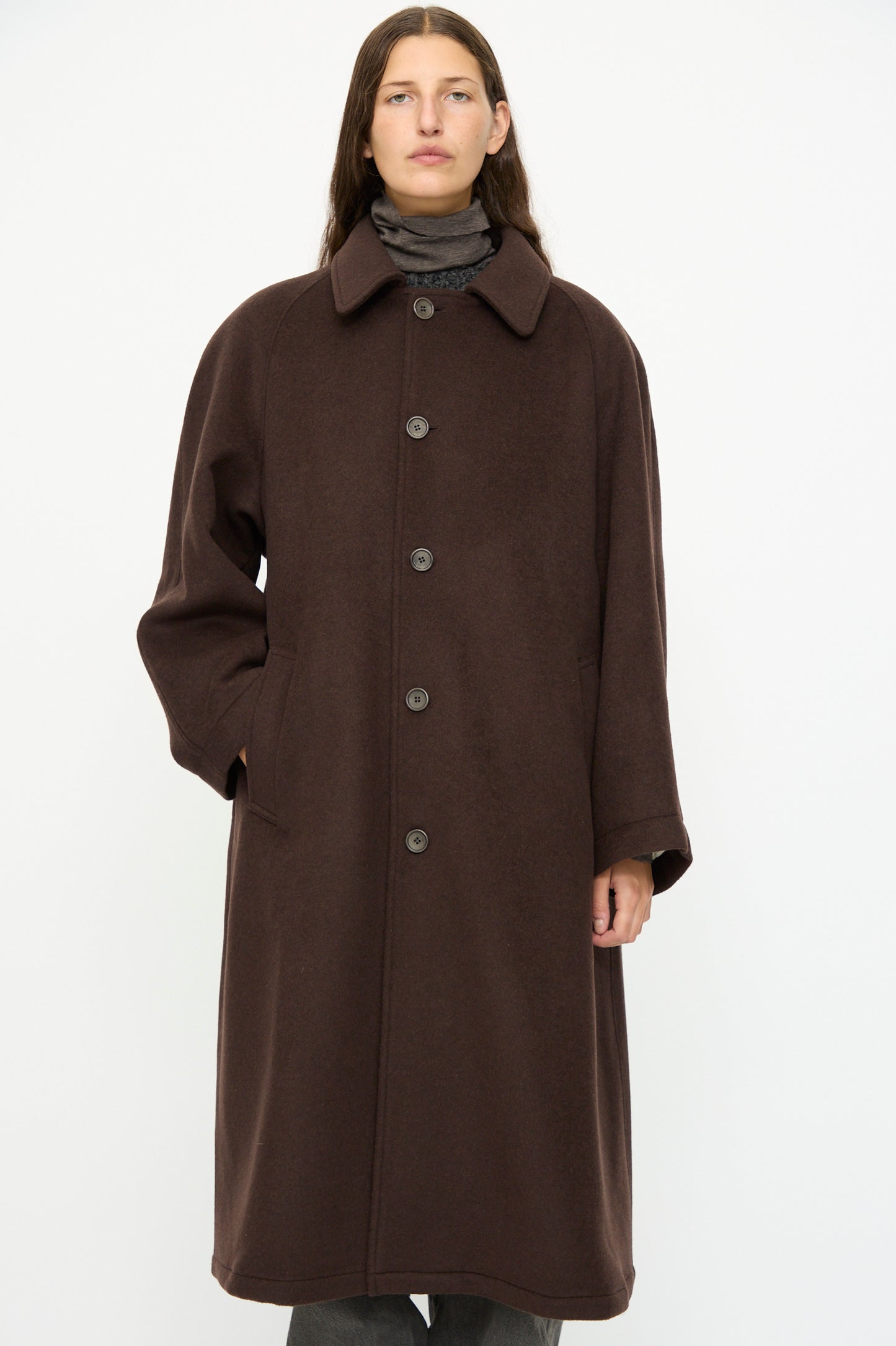 A person wearing the SONO Recycled Wool Carl Coat in Earth, an oversized dark brown coat with a collar and buttons, stands against a plain white background.