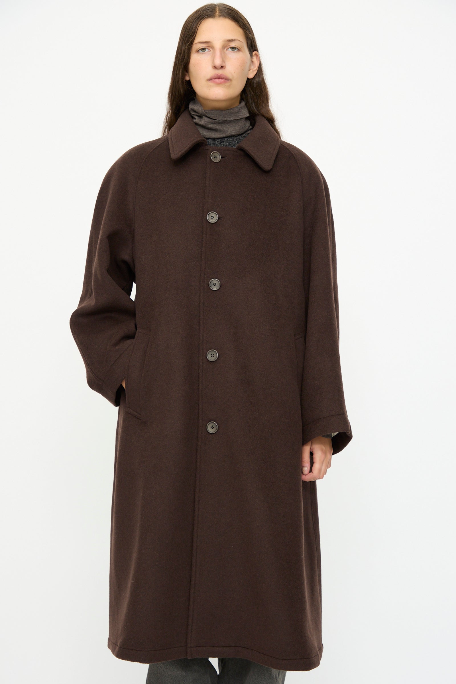 A person wearing the SONO Recycled Wool Carl Coat in Earth, an oversized dark brown coat with a collar and buttons, stands against a plain white background.
